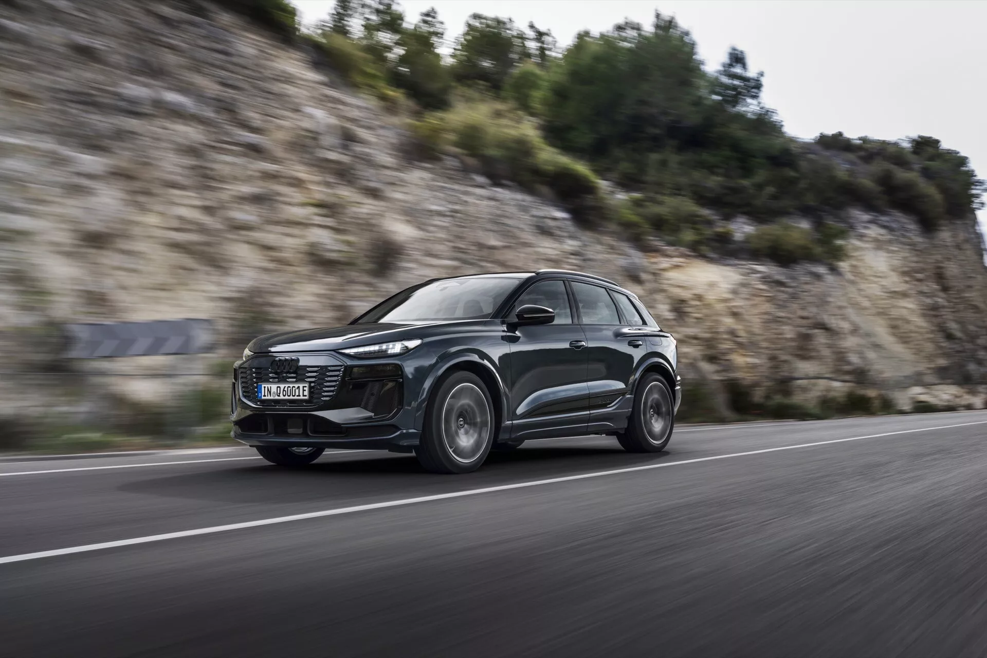 Prise dynamique de l'Audi Q6 e-tron 2024 en mouvement sous un angle bas, de face, mettant en valeur son design aérodynamique de SUV, ses phares LED modernes et ses roues en alliage avec flou de mouvement, suggérant des capacités de performance sur une route en asphalte.