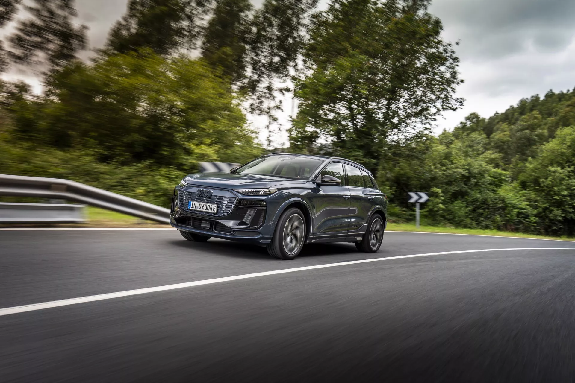 L'image montre un SUV Audi Q6 e-tron 2024 moderne en mouvement, capturé sous un angle avant latéral, se concentrant sur son côté gauche. La prise de vue dynamique met en évidence le phare avant gauche proéminent avec des feux de jour LED distinctifs, une grande calandre contemporaine, des rétroviseurs aérodynamiques avec clignotants intégrés et de grandes jantes en alliage avec des pneus à profil bas. La voiture dispose d'un toit panoramique et de vitres teintées, avec une peinture métallique réfléchissante. Le point de vue bas et le léger flou de l'arrière-plan soulignent la vitesse et le mouvement de la voiture sur une route à deux voies et courbes.