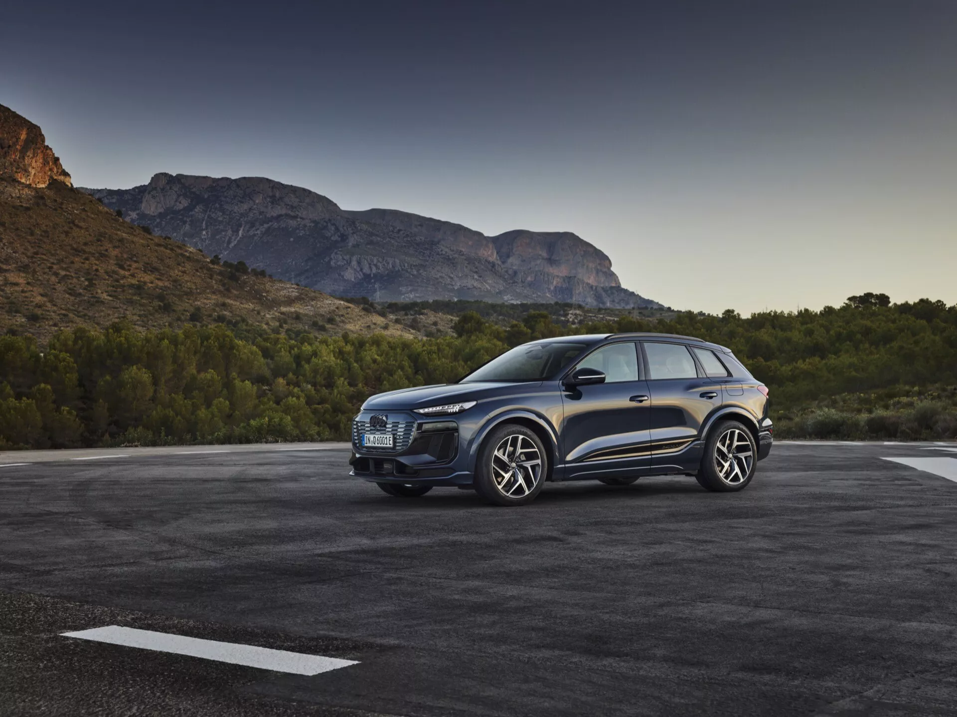 Vue de trois quarts avant d'un SUV bleu Audi Q6 e-tron 2024 garé en extérieur, mettant en évidence la calandre proéminente, les phares élégants, le capot sculpté, les grandes roues en alliage, les rétroviseurs latéraux et les portes du côté conducteur, le tout dans un décor de chaîne de montagnes sous la lumière du matin ou de fin d'après-midi.