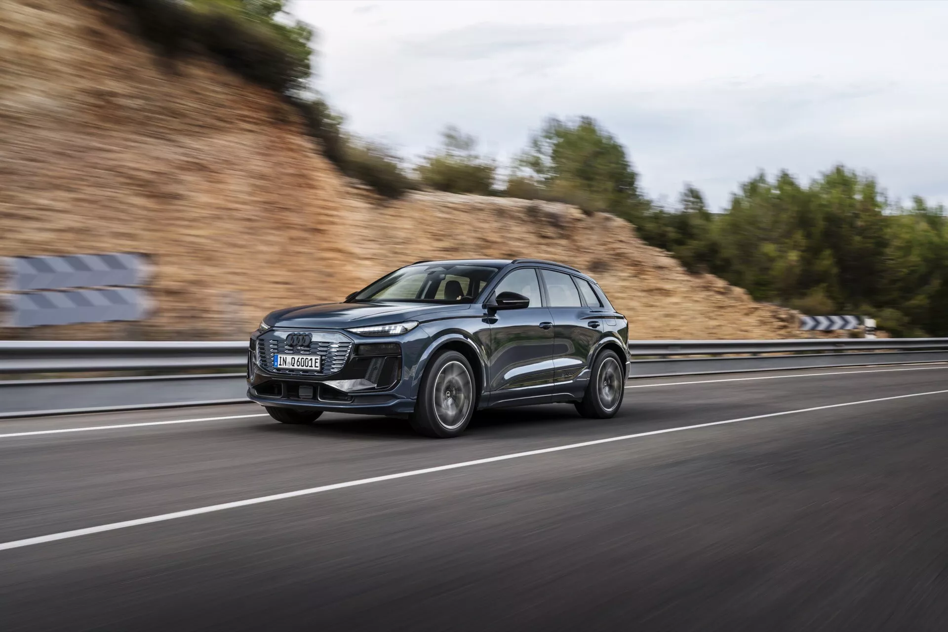 Image de l'Audi Q6 e-tron 2024 en mouvement, capturée à l'aide d'une technique de filé pour mettre en valeur le profil élégant et le design moderne du SUV. La voiture, cadrée sous un angle latéral, file sur une autoroute à plusieurs voies avec des roues et un arrière-plan flous, soulignant ses capacités dynamiques. Le véhicule de couleur métallique sombre est doté d'une grande calandre avant, de phares à LED distinctifs, de jantes en alliage sport et de surfaces réfléchissantes. Le ciel couvert et la route mouillée créent une scène dramatique, soulignant les caractéristiques de luxe et de performance de la voiture.
