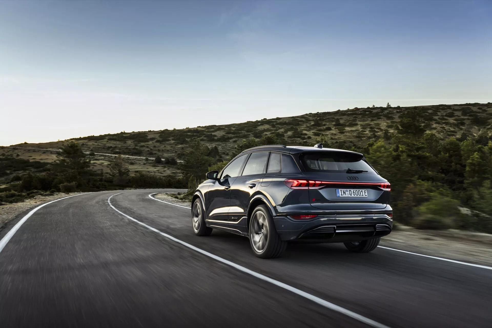 L'image capture une Audi Q6 e-tron 2024 sous un angle de trois quarts arrière, mettant en évidence le côté droit et l'arrière du véhicule. La voiture, apparaissant en mouvement sur une route sinueuse, présente ses éléments de design, y compris les feux arrière, le pare-chocs arrière, les sorties d'échappement, les jantes en alliage et une partie du profil latéral avec la porte arrière et la vitre passager. Le flou de l'arrière-plan implique la vitesse, et l'éclairage naturel suggère une prise de vue en journée, dans un environnement rural ou semi-rural avec une végétation clairsemée. Des caractéristiques avancées comme des feux arrière à LED, un becquet arrière ou des doubles échappements sont suggérées, bien que les détails technologiques plus fins soient indiscernables en raison du flou de mouvement.
