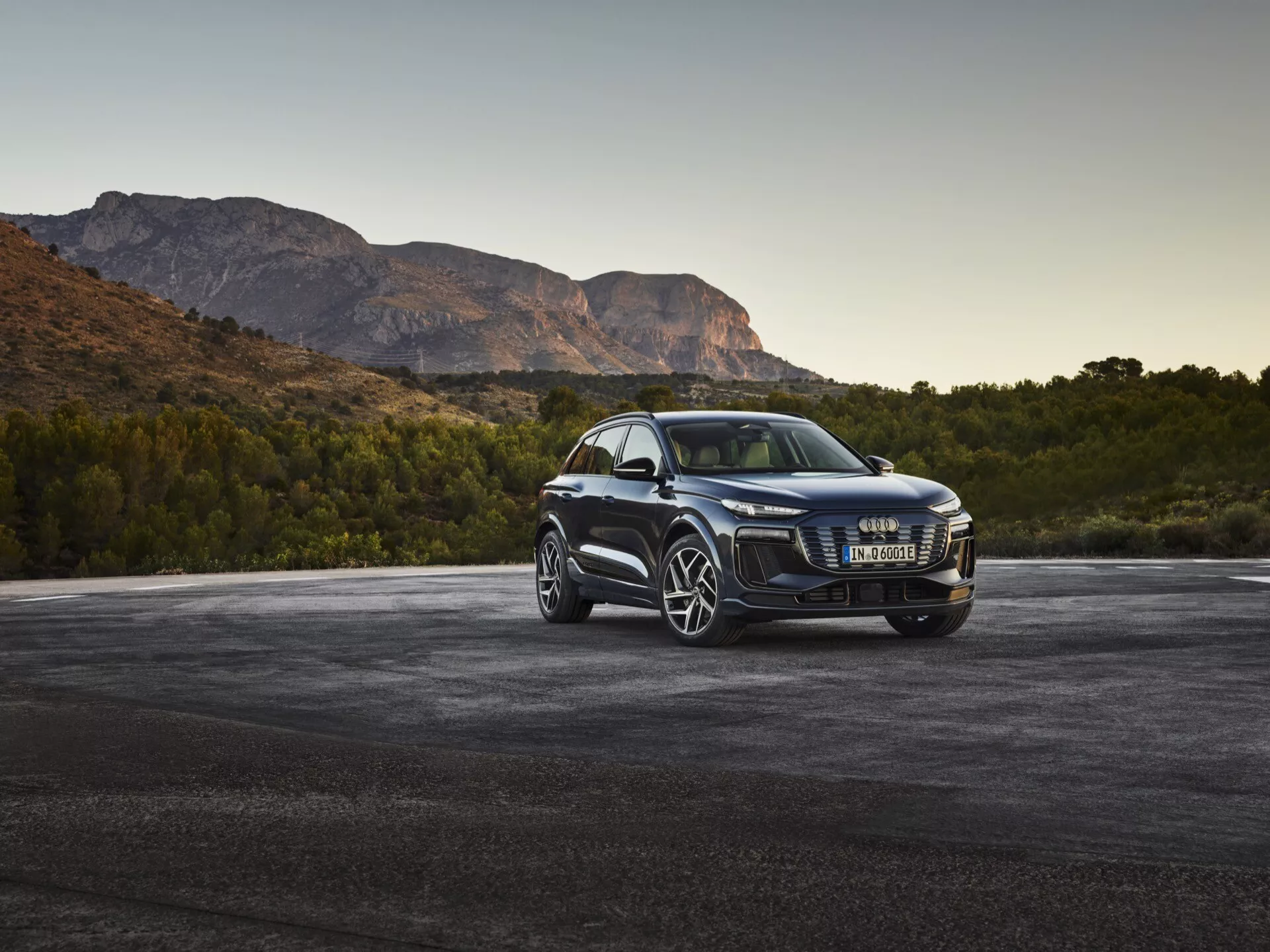 SUV de luxe Audi Q6 e-tron 2024 en angle avant de trois quarts montrant une grille entourée de chrome, des phares LED modernes, de grandes roues en alliage à plusieurs rayons, des rétroviseurs avec clignotants intégrés, des vitres teintées et une finition de peinture brillante, garé sur un asphalte avec des montagnes en arrière-plan au lever ou au coucher du soleil.