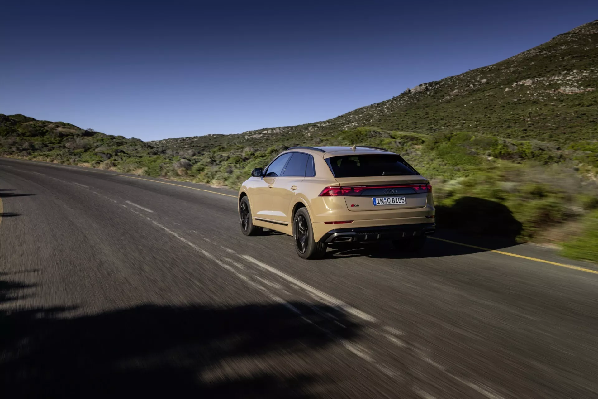 Audi Q8 2023 (4M-Facelift) en mouvement, capturée depuis un point de vue arrière trois quarts montrant le côté droit et arrière sur une route divisée avec un fond de collines naturelles sous un ciel bleu, présentant une ligne de toit type coupé, un design élégant, des feux arrière allongés, une peinture beige clair et un système d'échappement double.