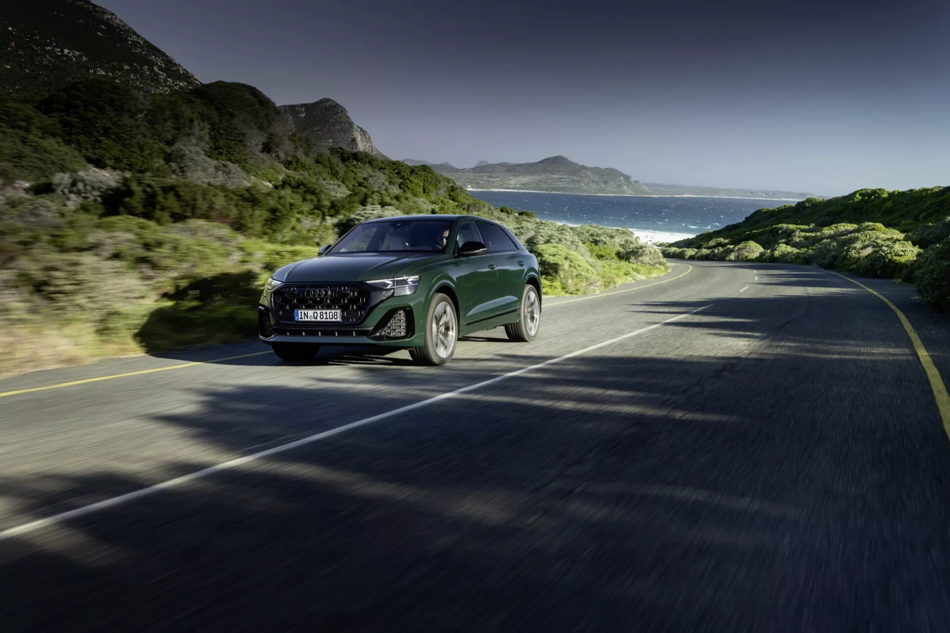 Audi Q8 2023 (4M-Facelift) roulant sur une route côtière, présentant une vue de face et de côté de sa grande grille, phares LED, capot élégant et panneau latéral partiel, incarnant la technologie et le luxe en mouvement contre un décor de ciel clair, de montagnes et d'océan.