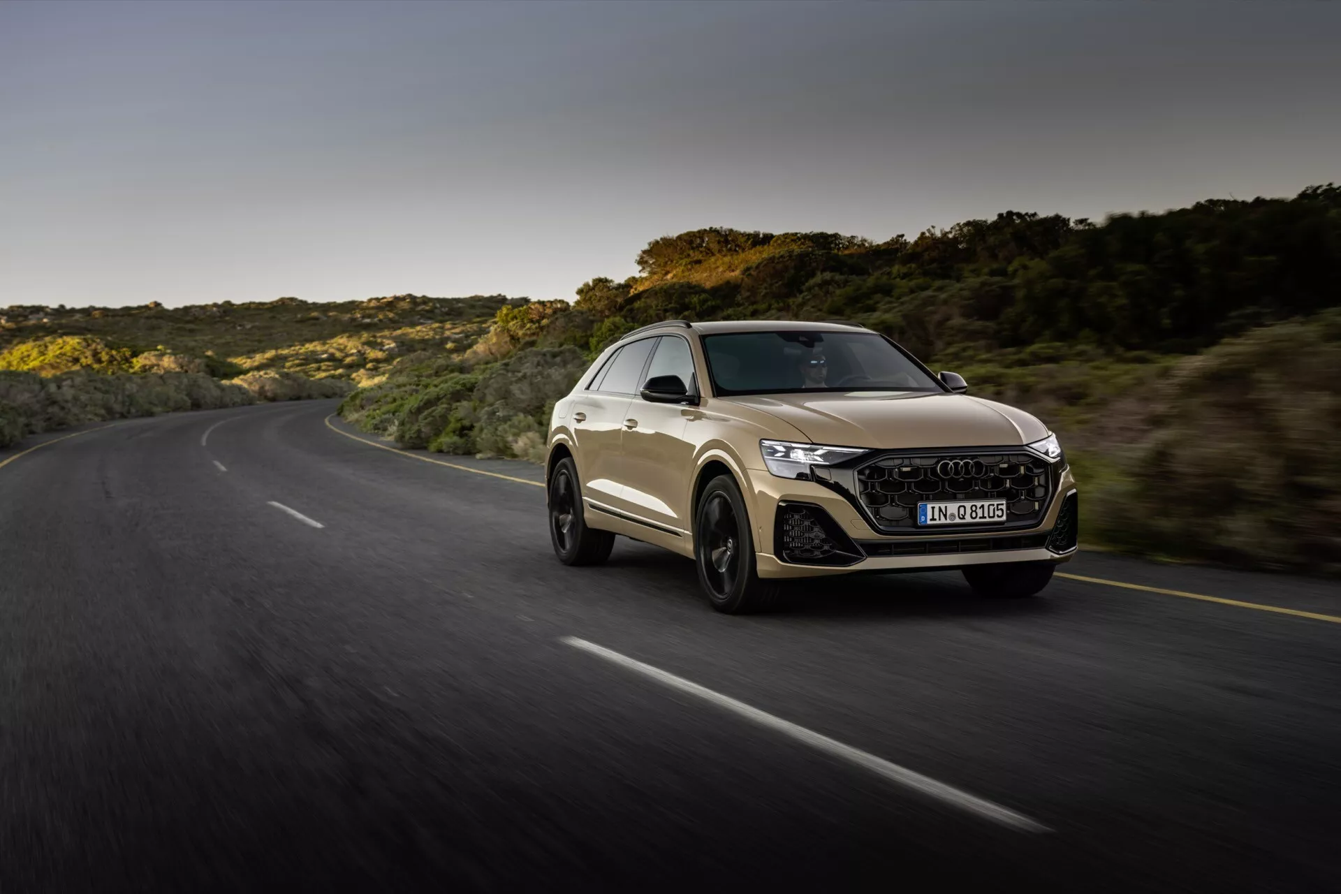 Audi Q8 2023 (4M-Facelift) en conduite dynamique sur une route sinueuse pendant l'heure dorée, mettant en avant le design sportif coupé de la voiture, l'avant agressif avec phares LED et grille hexagonale, et silhouette aérodynamique contre un fond de paysage naturel.