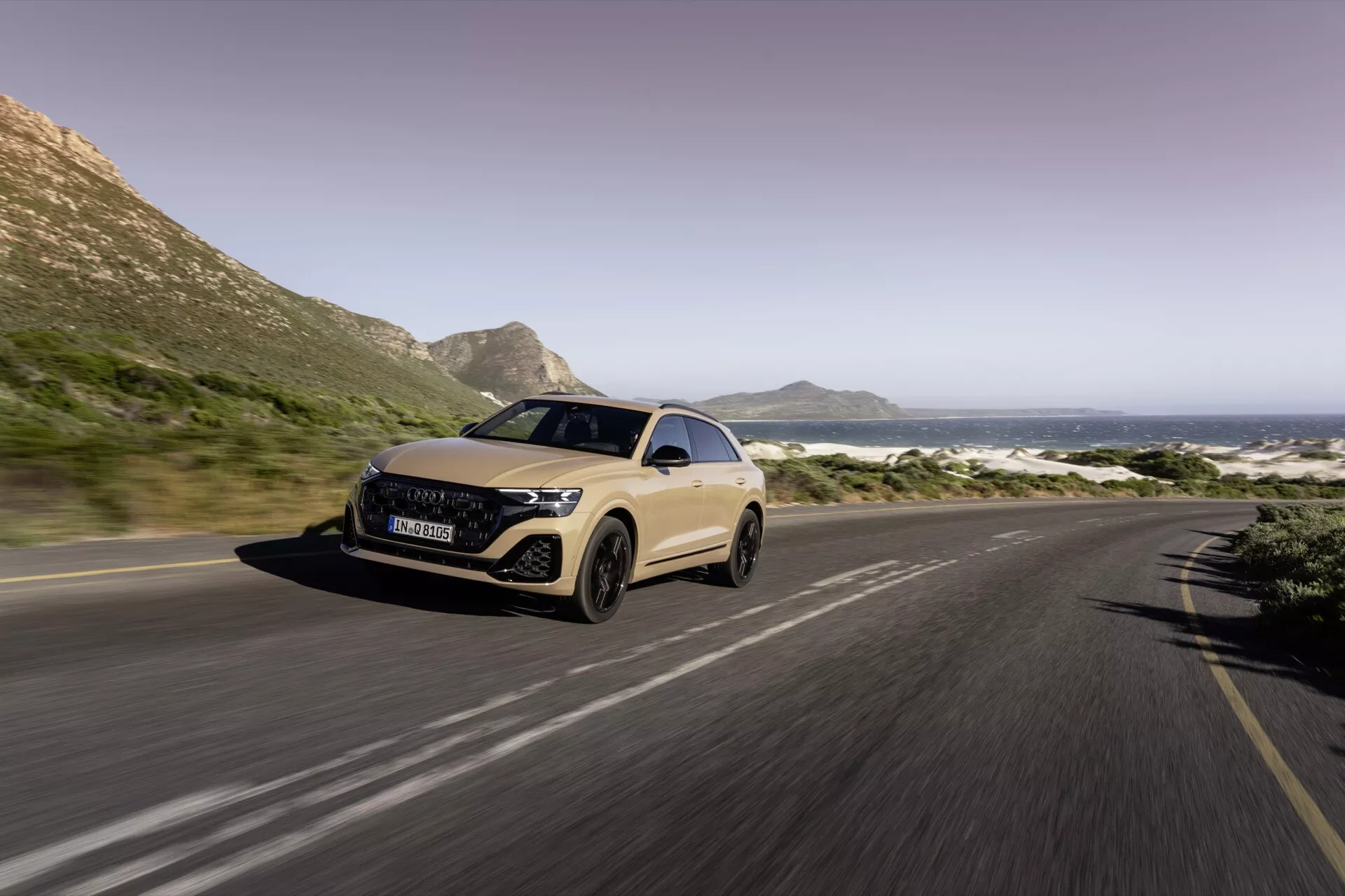 Vue de trois quarts avant de l'Audi Q8 2023 (4M-Facelift) sur une route côtière, montrant les phares LED, la conception aérodynamique et le flou de mouvement indiquant le mouvement