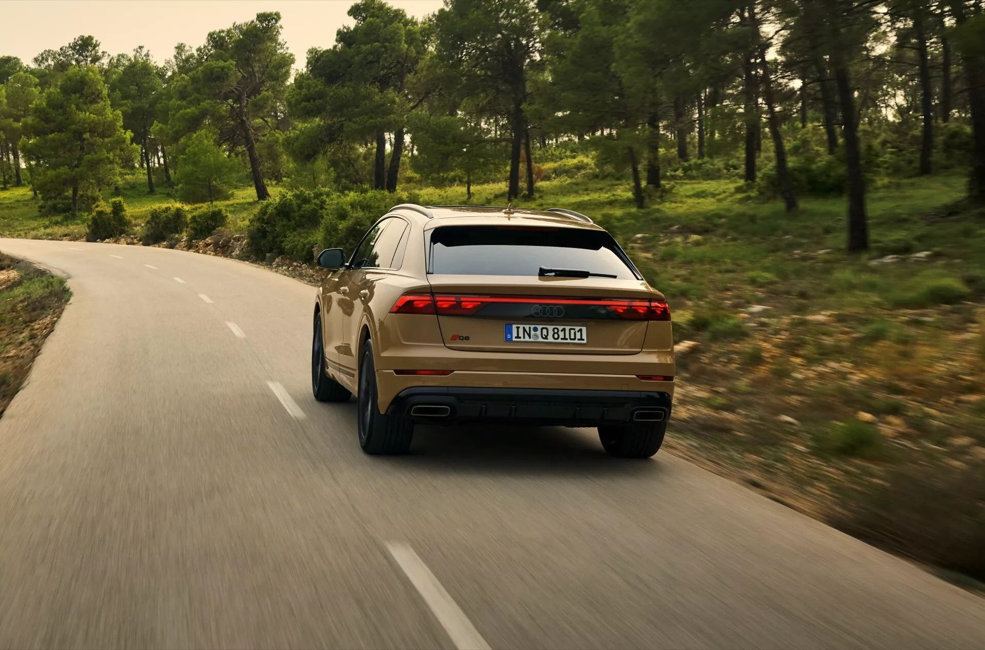 Vue arrière d'une Audi Q8 2023 (4M-Facelift) circulant sur une route sinueuse à travers une zone boisée, montrant le quart arrière droit avec feux arrière LED, double échappement et caractéristiques de conception moderne