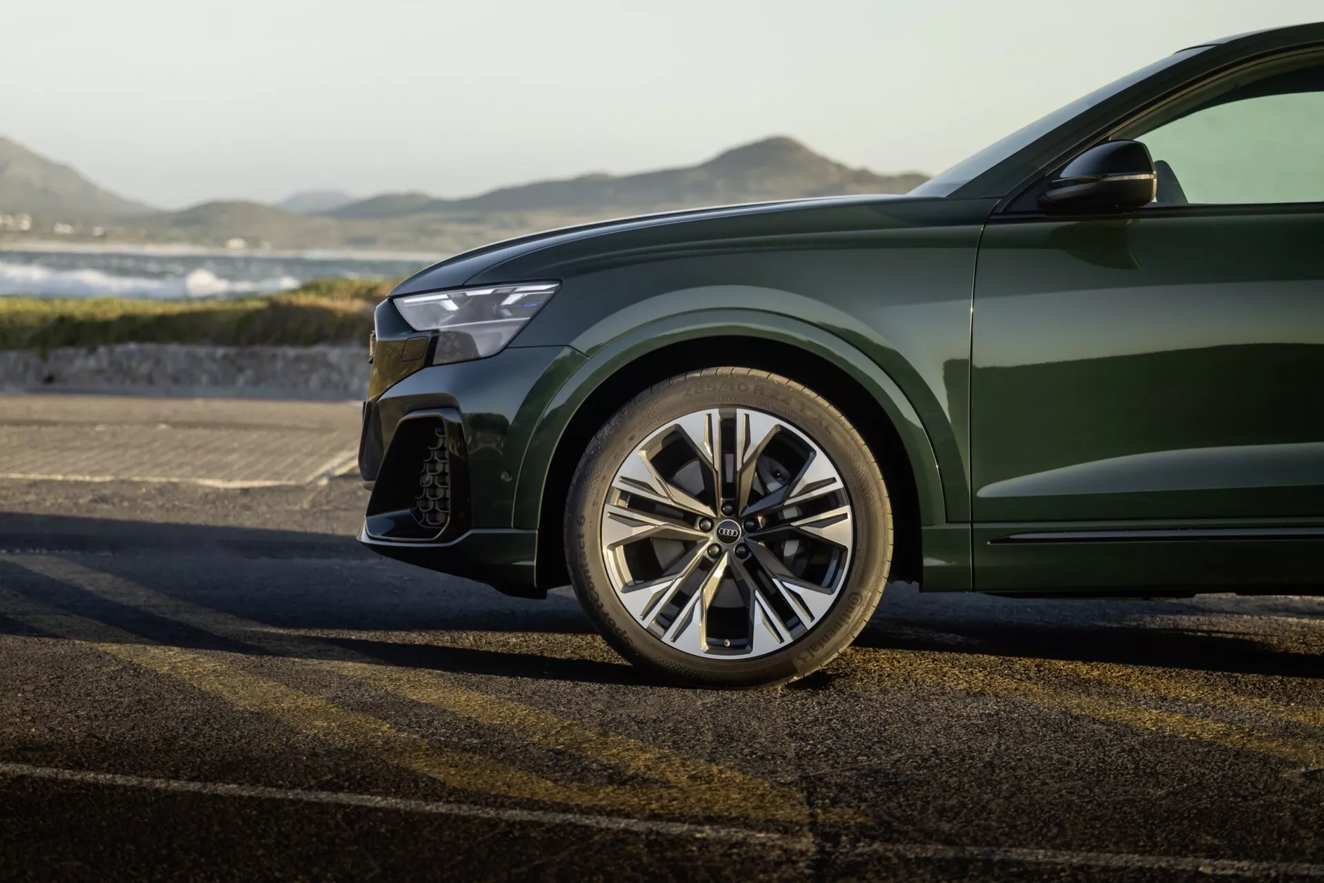 Vue rapprochée du quart avant gauche sportif de l'Audi Q8 2023 (4M-Facelift), montrant la roue en alliage à multi-rayons polie, portion du pare-chocs avant avec grande prise d'air, bord du phare LED moderne et peinture métallique vert foncé brillante sur la carrosserie, sur fond extérieur.