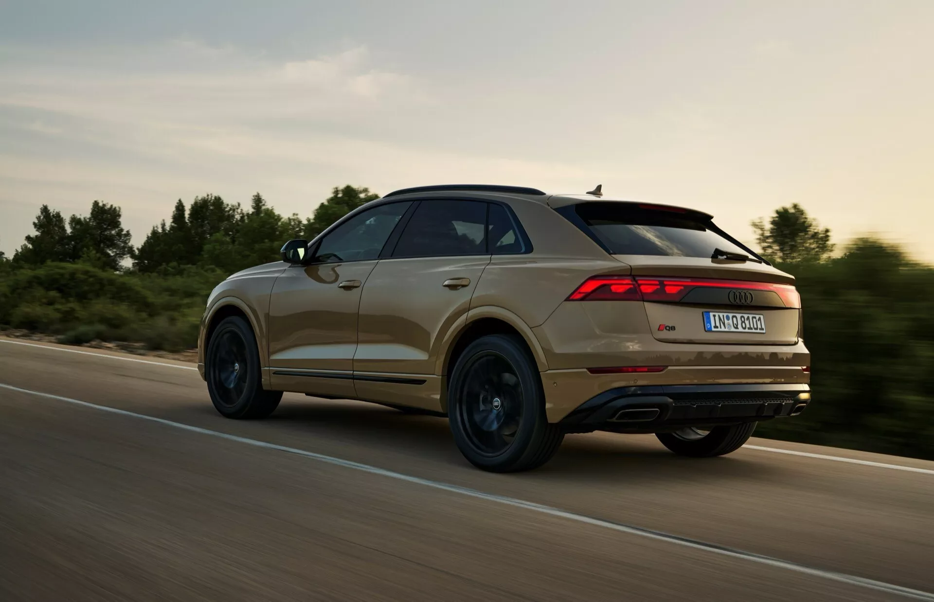 Vue arrière trois quarts d'un SUV Audi Q8 (4M-Facelift) 2023 en mouvement, présentant un design distinctif des feux arrière, des lignes de contour et le profil des roues, capturée lors d'une session de conduite dynamique dans un cadre rural avec un éclairage chaleureux.