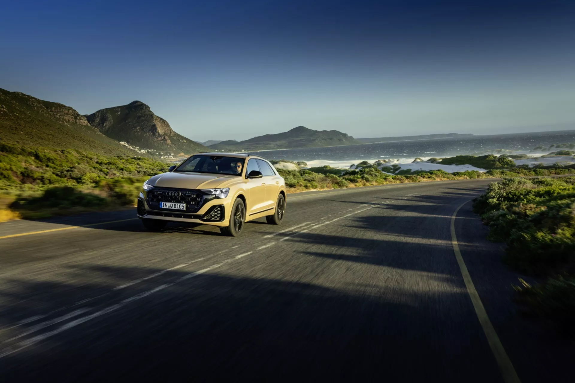 Audi Q8 2023 (4M-Facelift) conduisant le long d'une route côtière, exposant son design dynamique et ses phares LED dans une vue de trois quarts avant avec flou de mouvement