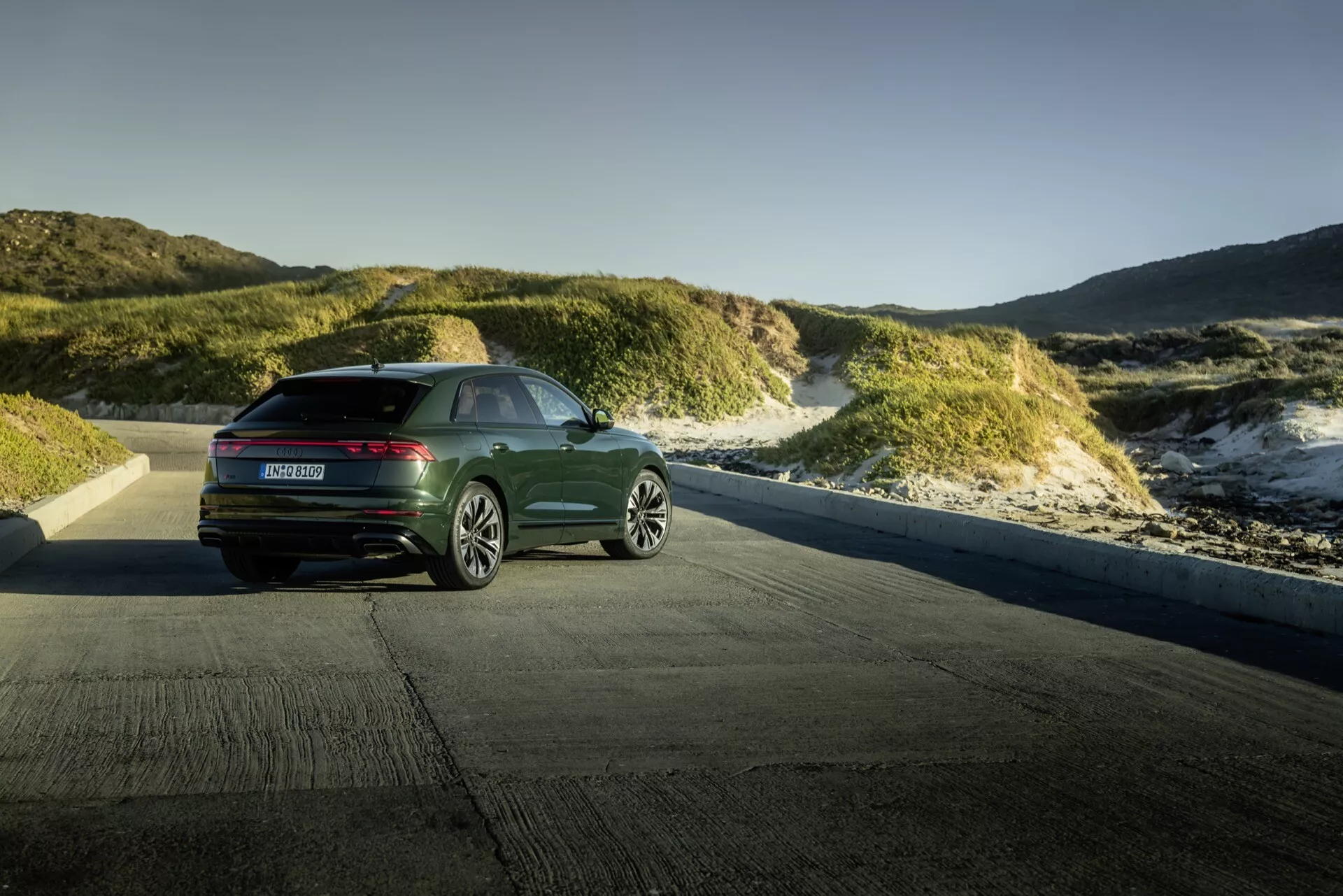 Audi Q8 4M-Facelift 2023 présentée contre un paysage de dunes côtières à l'heure dorée, montrant une vue arrière trois quarts avec des feux arrière horizontaux modernes et un design élégant