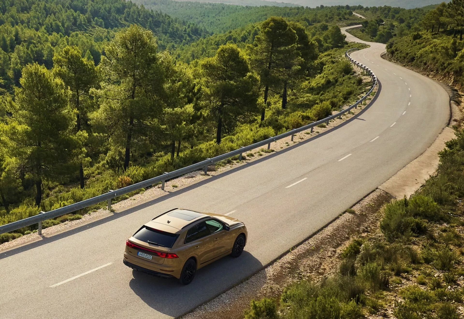 Audi Q8 2023 (4M-Facelift) couleur or sur une route sinueuse à deux voies dans un environnement rural ou forestier, vue sous un angle surélevé montrant son design arrière et latéral contre le paysage naturel