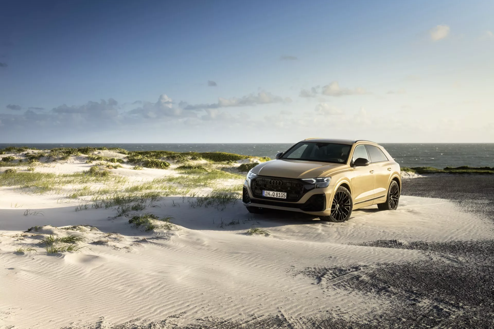 Audi Q8 2023 (4M-Facelift) stationnée sur un terrain sablonneux sous un ciel partiellement nuageux avec la mer en arrière-plan, mettant en avant la grille avant, les phares LED et le design aérodynamique