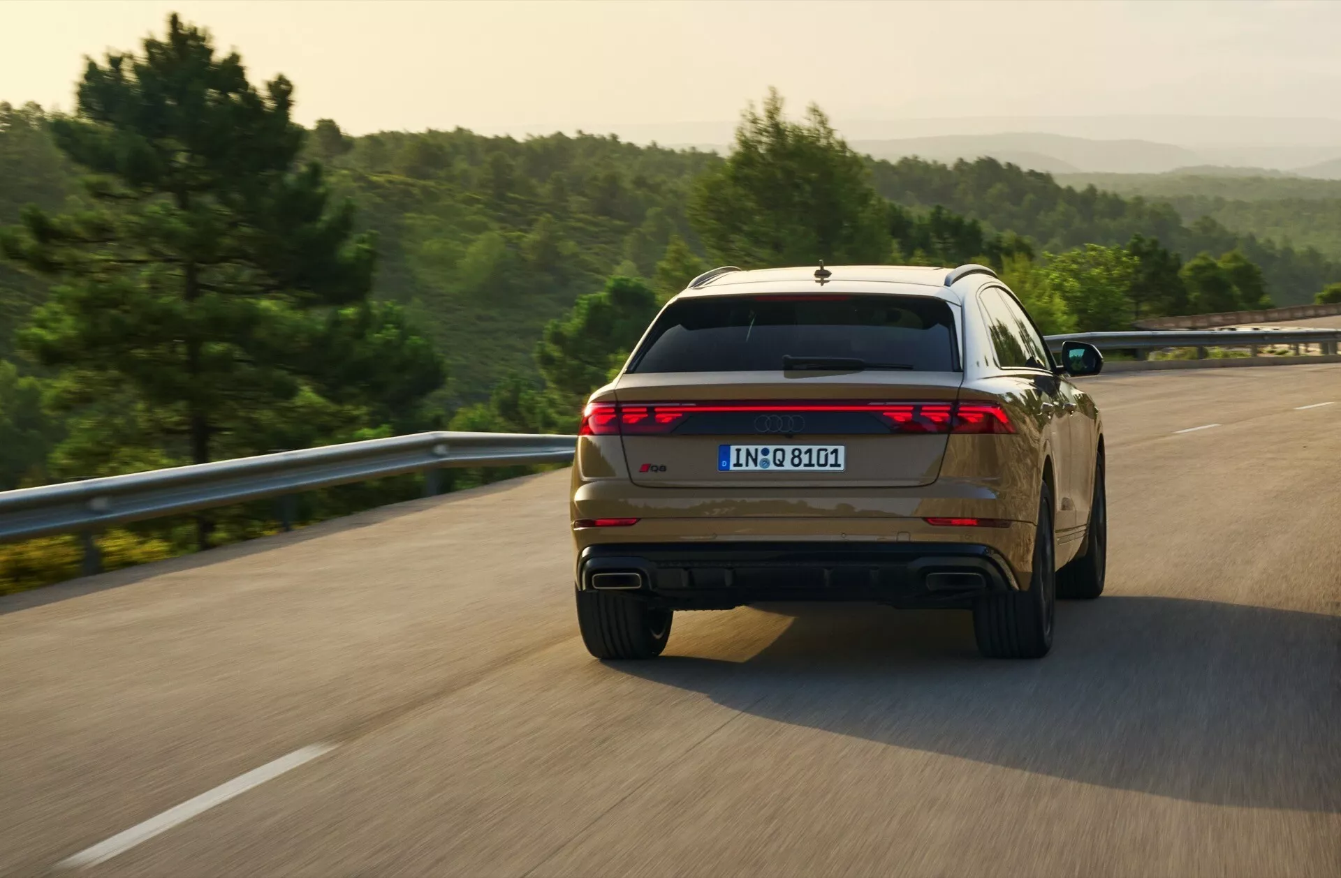 Audi Q8 2023 (4M-Facelift) capturé dans une vue arrière dynamique de trois quarts lors de la conduite sur une route ouverte, présentant son design de feux arrière sur toute la largeur et des embouts d'échappement doubles