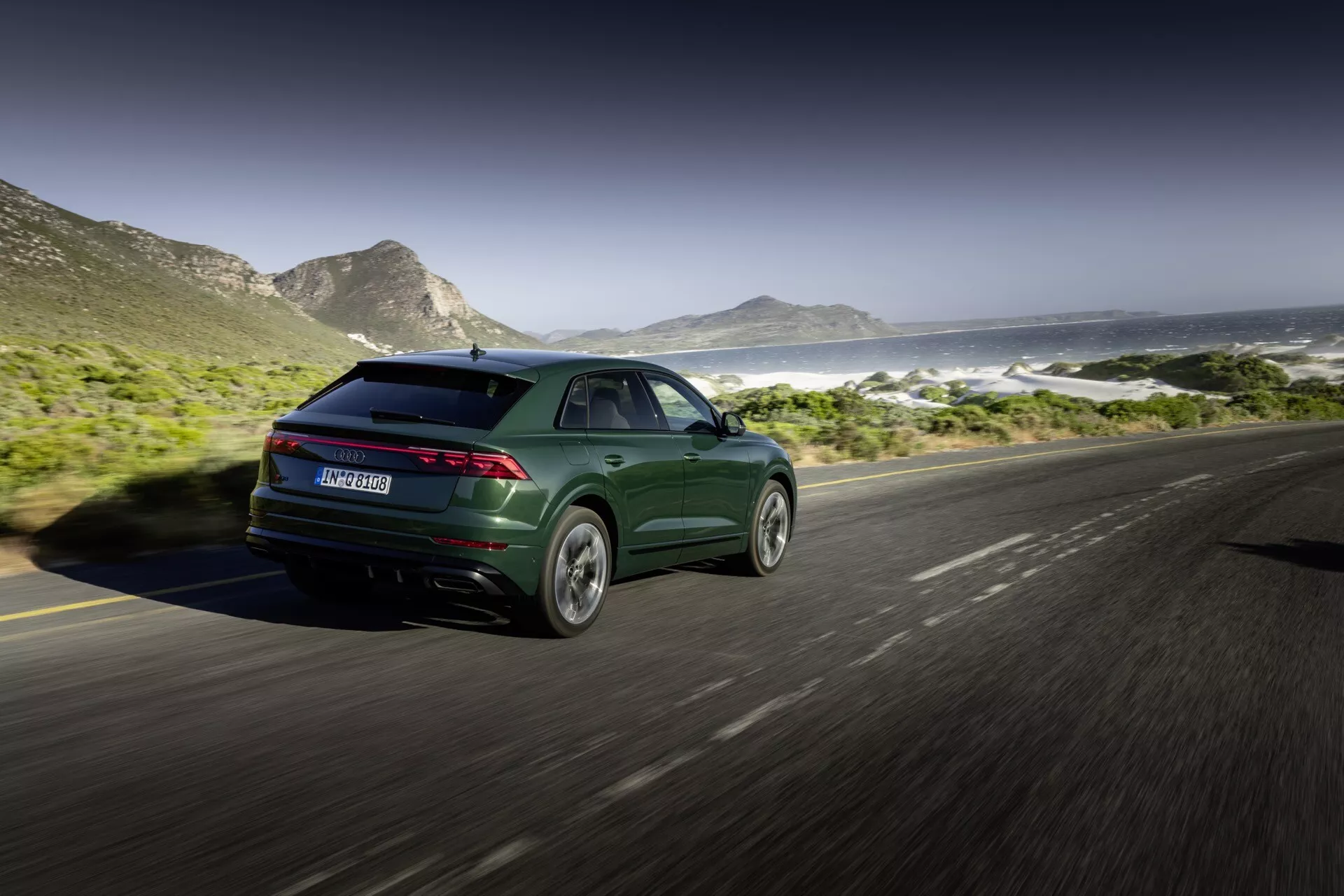 Audi Q8 2023 (4M-Facelift) en vert foncé métallisé, présentant son design sportif et moderne avec des feux arrière élancés et des jantes en alliage, capturé depuis un angle arrière de trois quarts sur une route côtière avec des montagnes et la mer en arrière-plan