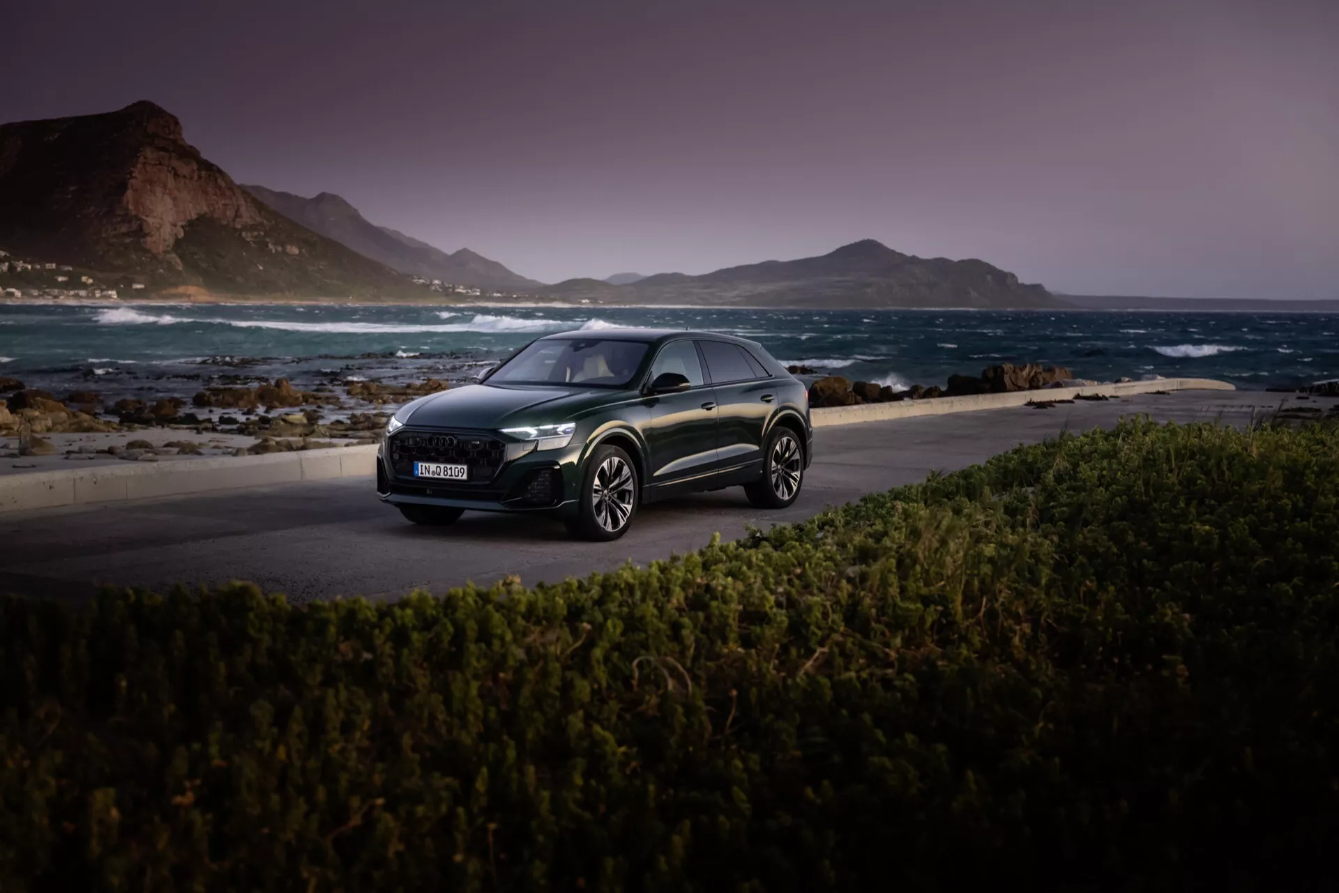 Audi Q8 2023 (4M-Facelift) hatchback de couleur sombre photographiée sur une route côtière pittoresque au crépuscule, mettant en avant ses phares LED, son profil aérodynamique et ses jantes en alliage