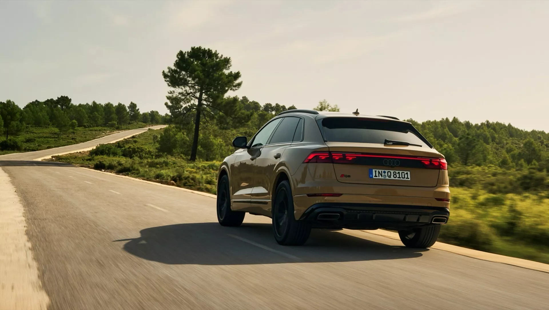 Audi Q8 2023 (4M-Facelift) en mouvement sur une route sinueuse, présentant ses profils arrière et latéral sportifs avec barre lumineuse LED, double échappement et finition métallique