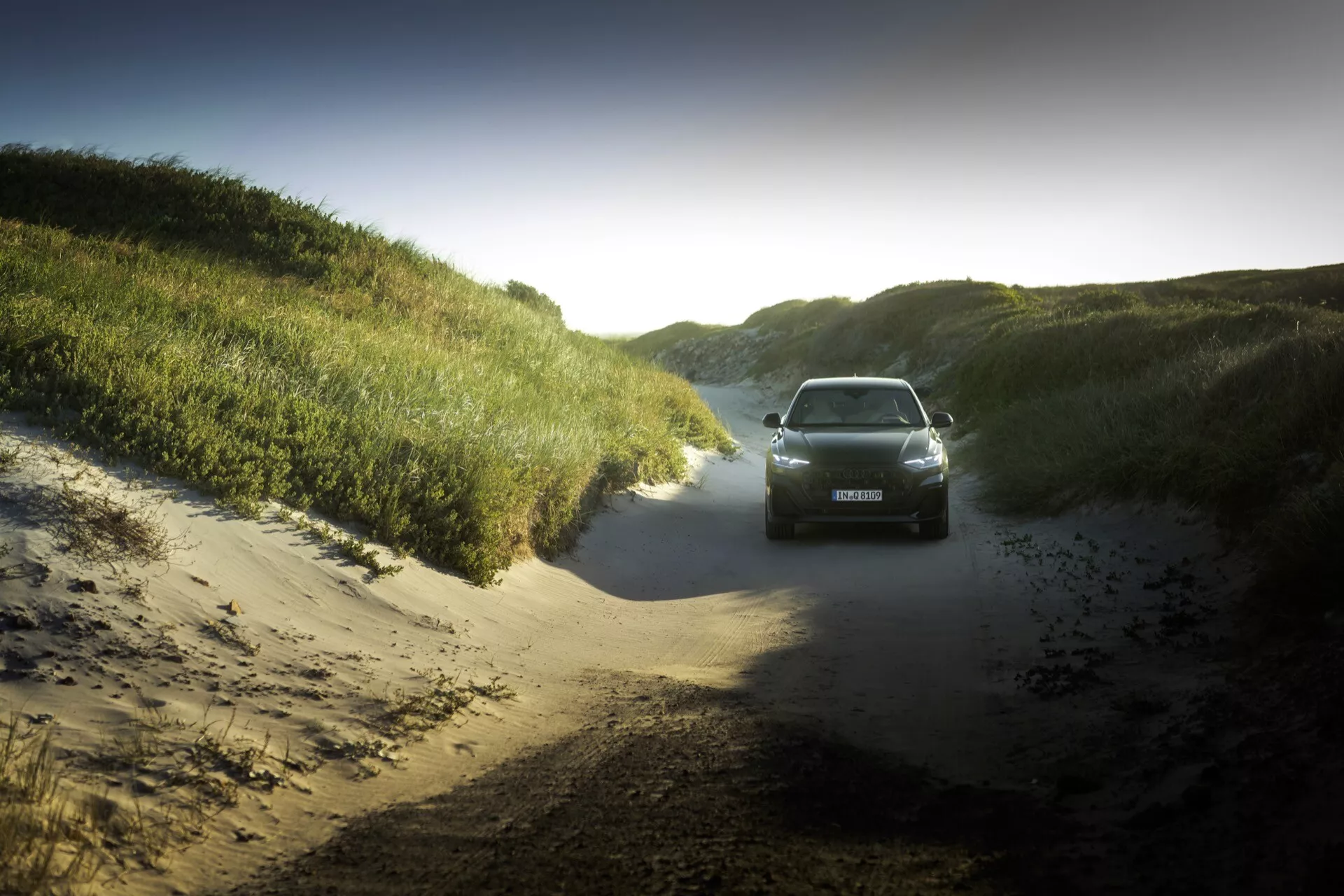 Audi Q8 2023 (4M-Facelift) capturée depuis une perspective frontale avec un léger angle élevé sur un chemin de terre, mettant en évidence son potentiel aventureux et son design élégant au milieu d'une silhouette dramatique au lever ou coucher du soleil