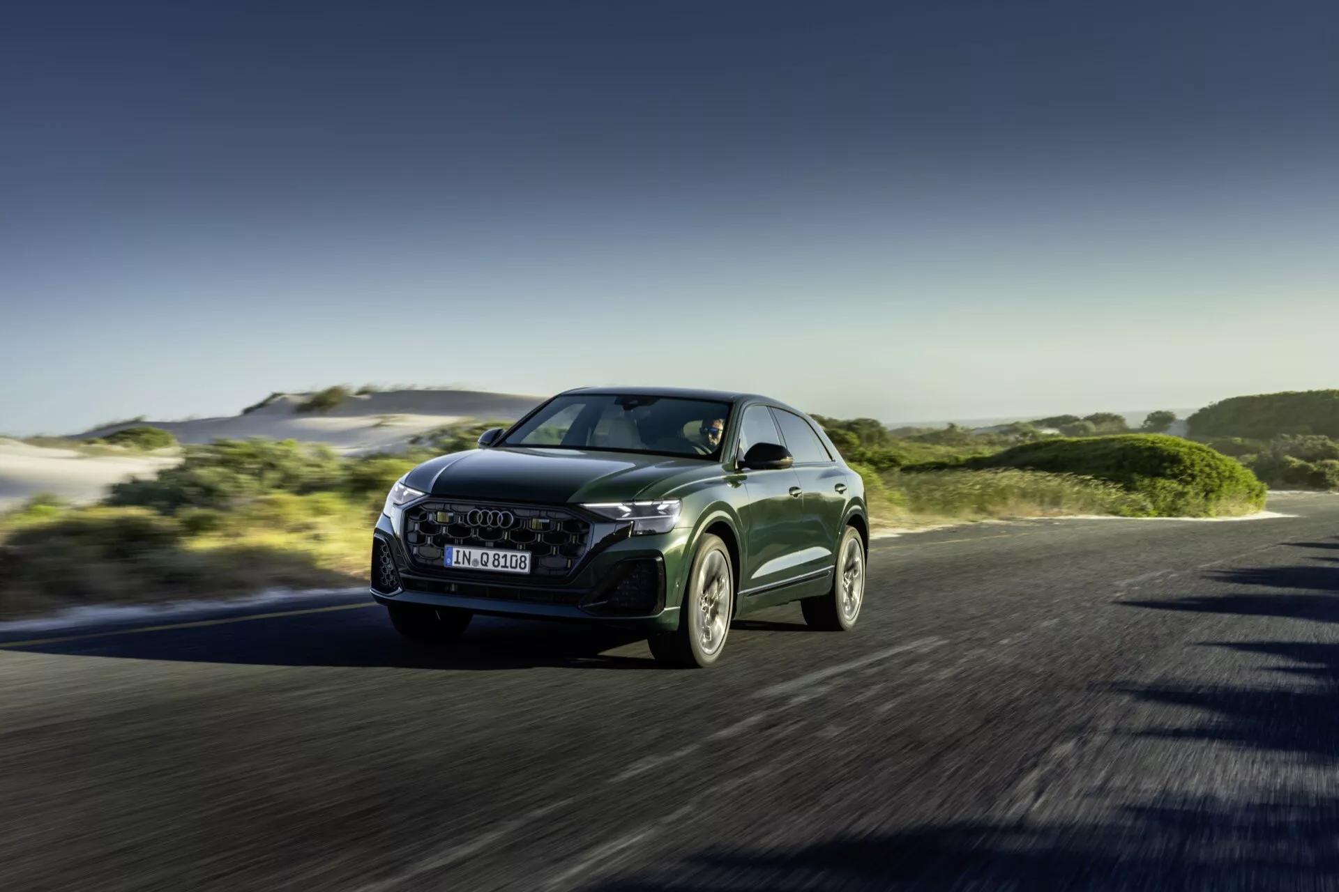 Audi Q8 2023 (4M-Facelift) en mouvement, présentant son design aérodynamique et élégant et ses phares LED depuis un angle dynamique bas avant-gauche, avec une vue trois quarts mettant en évidence la façade avant agressive du véhicule contre un arrière-plan de paysage naturel ensoleillé