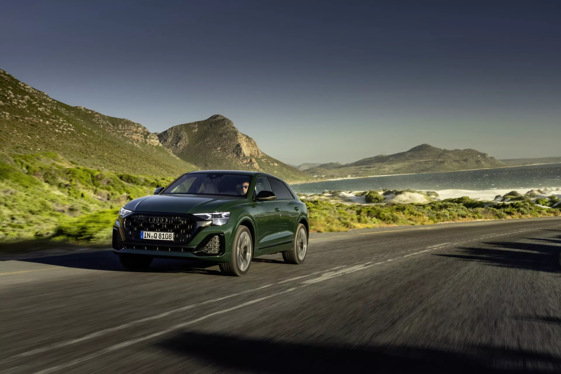 Audi Q8 4M Facelift 2023 roulant sur route côtière avec vue dynamique de trois quarts avant présentant des phares LED, calandre moderne et effet de flou de mouvement sur un fond d'océan et de montagnes sous un ciel ensoleillé