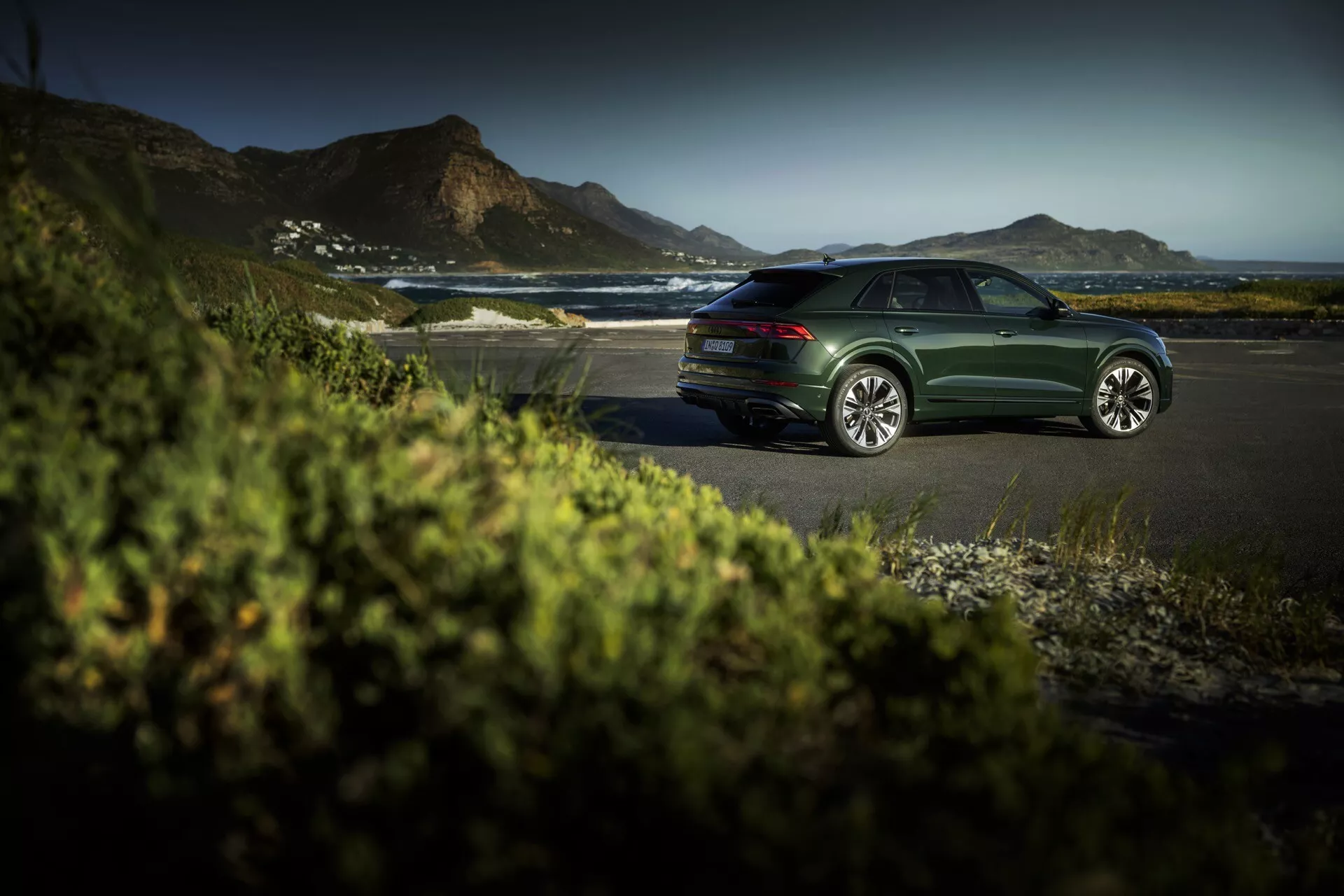 Sedan Audi Q8 (4M-Facelift) vert foncé garé sur une route côtière à l'heure dorée, présentant des lignes de design élégantes, des roues en alliage à multiples rayons et un cadre extérieur serein et luxueux