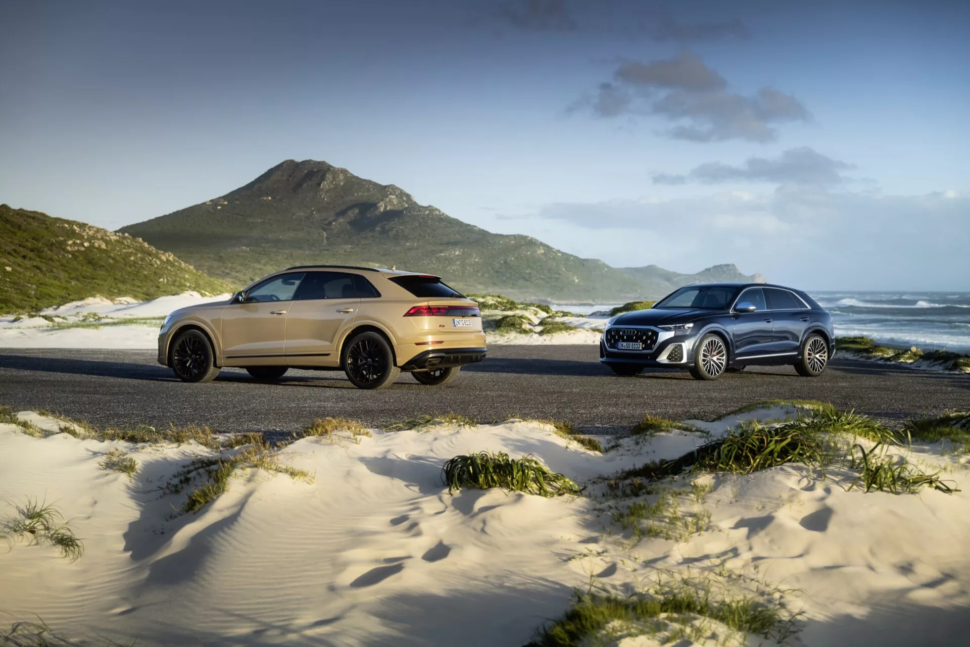 Modèles Audi Q8 (4M-facelift) de 2023 présentés sur une plage de sable, avec un SUV sportif doré à gauche en vue arrière de trois quarts et un SUV traditionnel argenté à droite en vue avant de trois quarts, situés contre des dunes herbeuses, des montagnes et un ciel partiellement nuageux