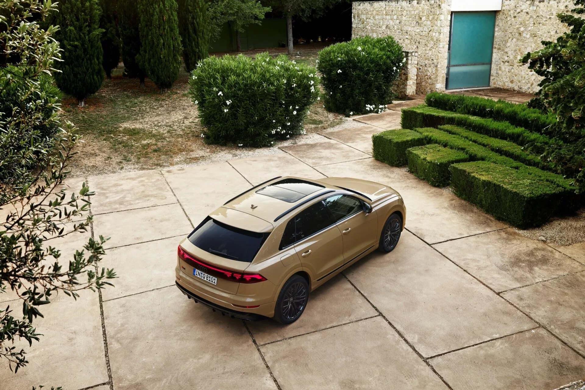 Berline Audi Q8 2023 (4M-facelift) vue de haut en arrière et de côté montrant des feux arrière LED, un toit ouvrant panoramique et des jantes sportives, garée avec un fond de haies soignées et un bâtiment en pierre