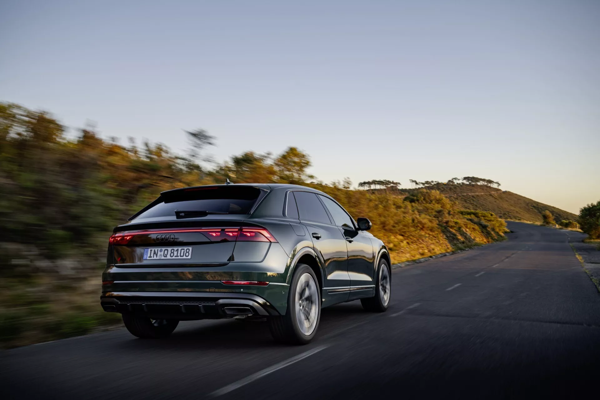 Audi Q8 2023 (4M-Facelift) en mouvement, capturée depuis un angle arrière de trois quarts montrant les caractéristiques de design moderne du véhicule incluant des feux arrières horizontaux élégants sur toute la largeur, des tuyaux d'échappement doubles, un diffuseur arrière et un spoiler intégré sur un fond de paysage naturel sur une route ouverte au coucher ou lever du soleil, soulignant son orientation performance et esthétique avancée.