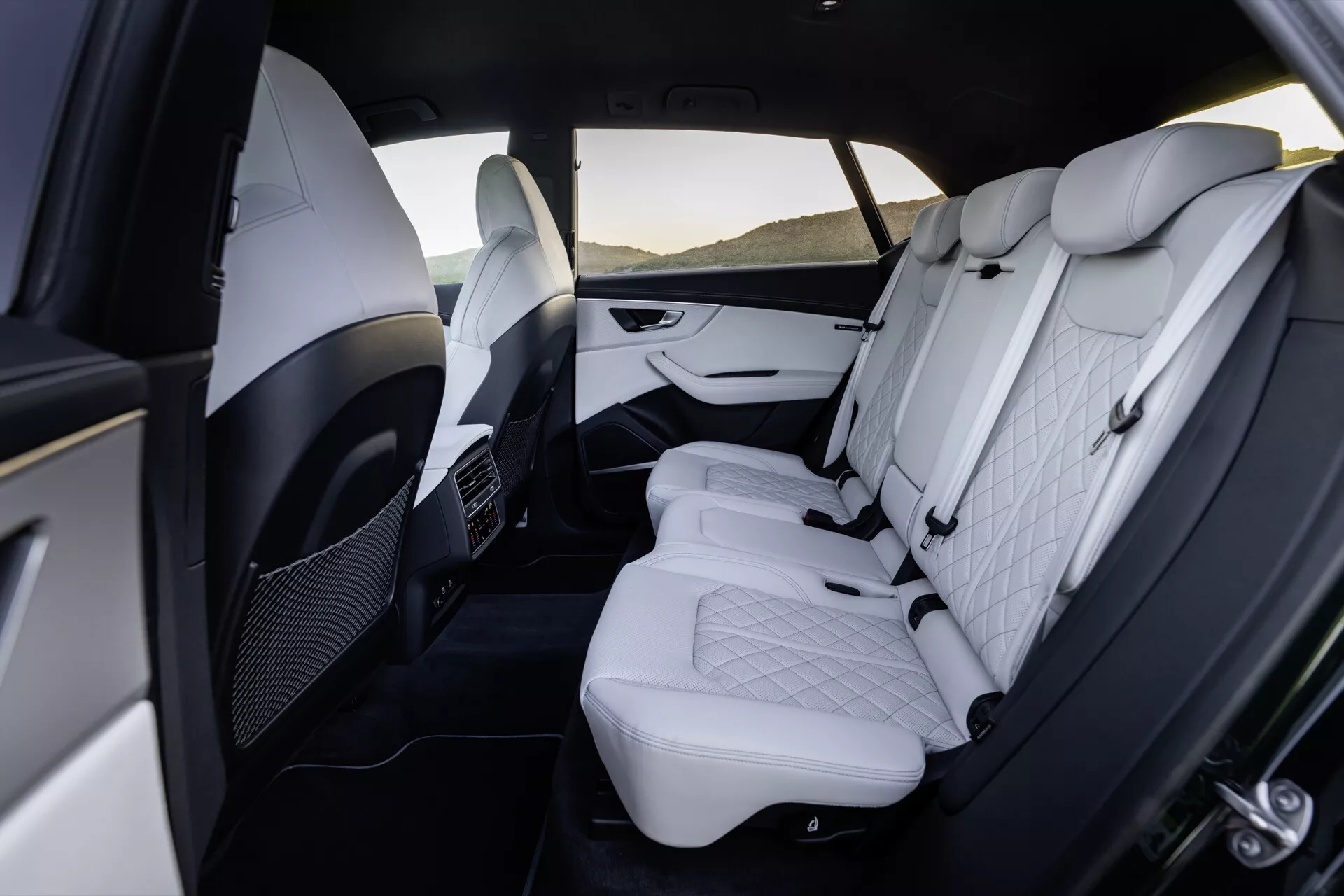 Vue du compartiment arrière intérieur de luxe de l'Audi Q8 2023 (4M Facelift), exposant un revêtement en cuir synthétique blanc premium avec motif losange matelassé, appuis-tête ajustables, panneaux de porte bicolores avec accents chromés, accoudoir rabattable, bouches d'aération pour les passagers arrière et espace généreux pour les jambes.