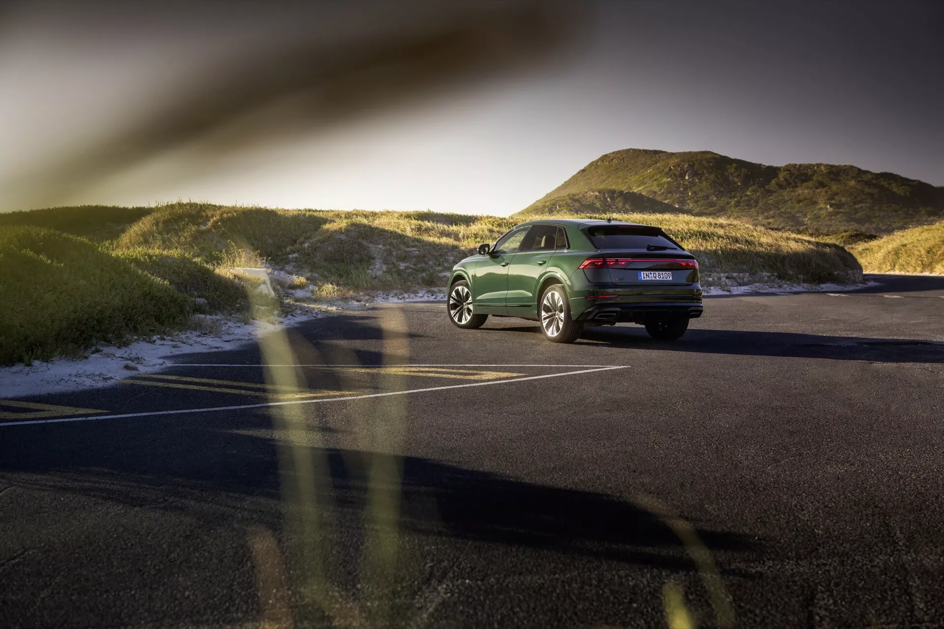 Vue arrière trois quarts de l'Audi Q8 (4M-Facelift) 2023 sur une aire de stationnement asphaltée au coucher du soleil, mettant en évidence sa posture, ses lignes de conception, ses feux arrière à LED, ses roues en alliage et sa finition verte polie avec un paysage naturel en arrière-plan.