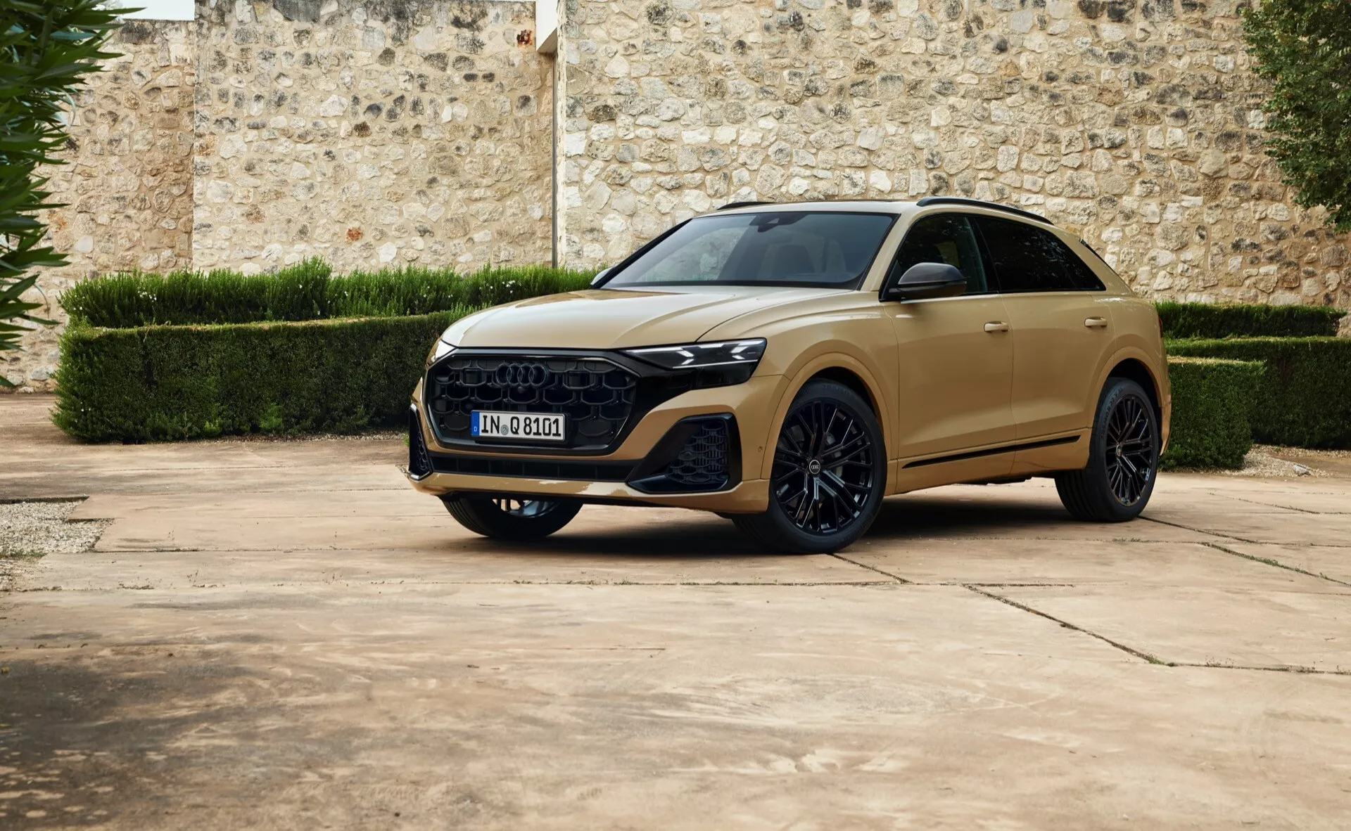 SUV crossover de luxe Audi Q8 2023 en vue de trois quarts avant stationné sur une surface pavée avec un fond de mur en pierre ancienne, montrant des phares LED élégants, une grille hexagonale proéminente, des lignes sculptées sur le capot, de grandes roues en alliage, et un possible toit panoramique.