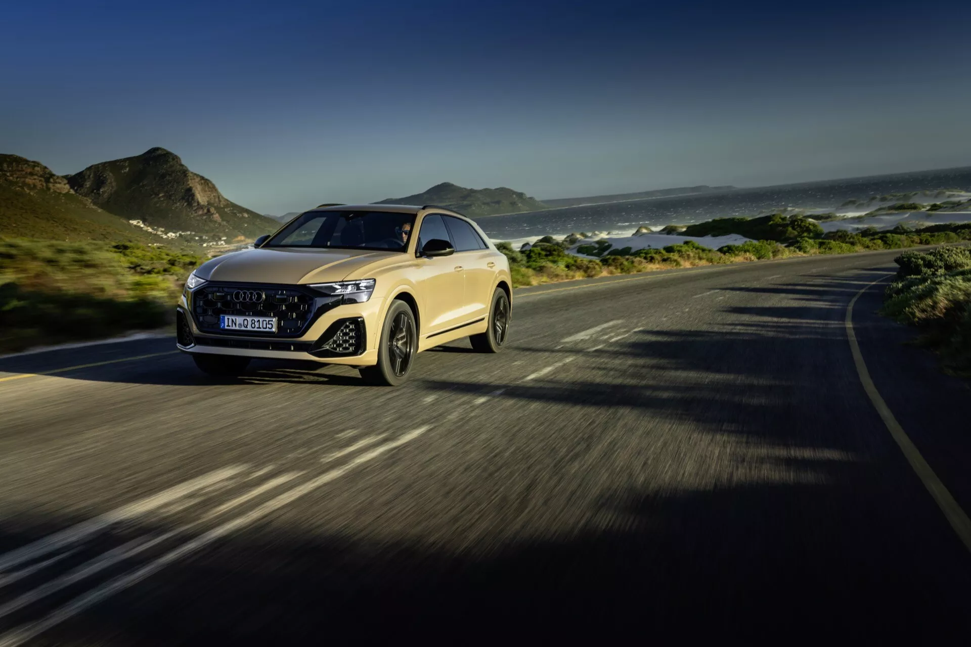 Audi Q8 4M facelift 2023 en mouvement sur une route côtière, mettant en évidence les phares LED avancés de la voiture de luxe, la calandre proéminente, le capot aérodynamique et la maniabilité dynamique, le tout sur un fond de montagnes et océan avec un accent sur les caractéristiques de design et technologie.