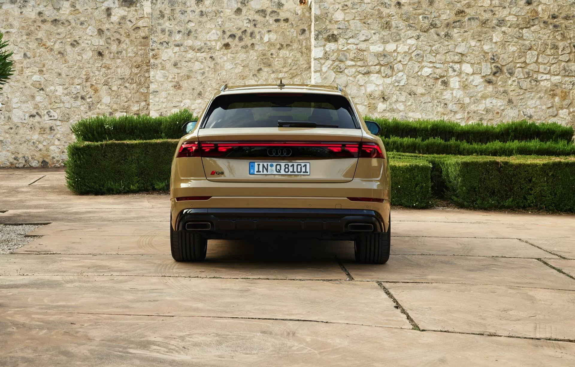 Audi Q8 2023 (4M-Facelift) présentant une vue arrière avec des feux arrière LED modernes, des échappements doubles, une plaque d'immatriculation européenne, stationnée sur une surface pavée contre un décor de mur en pierre et de haies vertes taillées, soulignant les caractéristiques de luxe et de design avancé