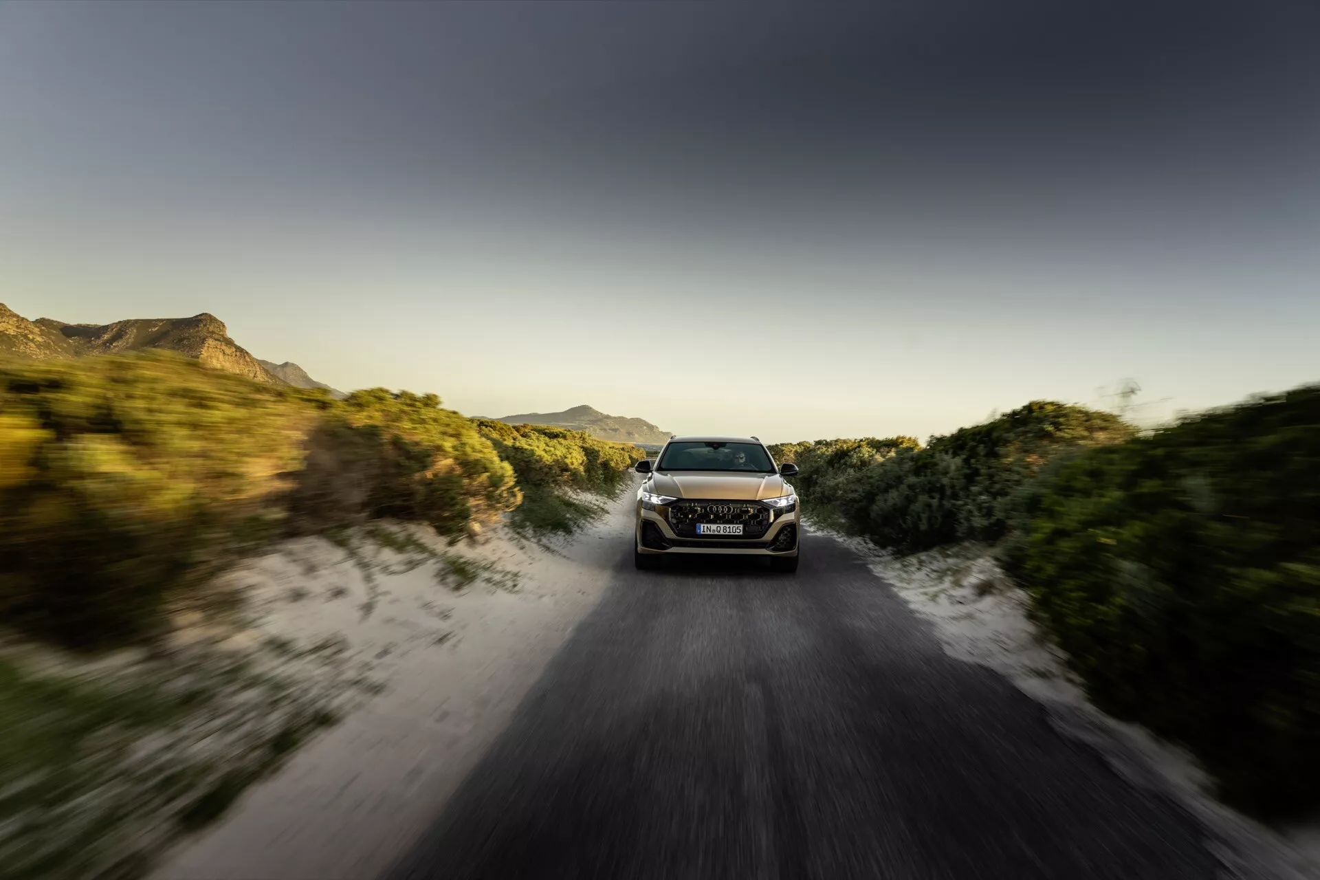 Audi Q8 2023 (4M-Facelift) en mouvement dynamique sur une route non pavée à l'aube ou au coucher du soleil avec effet de flou de mouvement, mettant en avant ses phares LED, design aérodynamique élégant et capacité tout-terrain au milieu d'un cadre rural