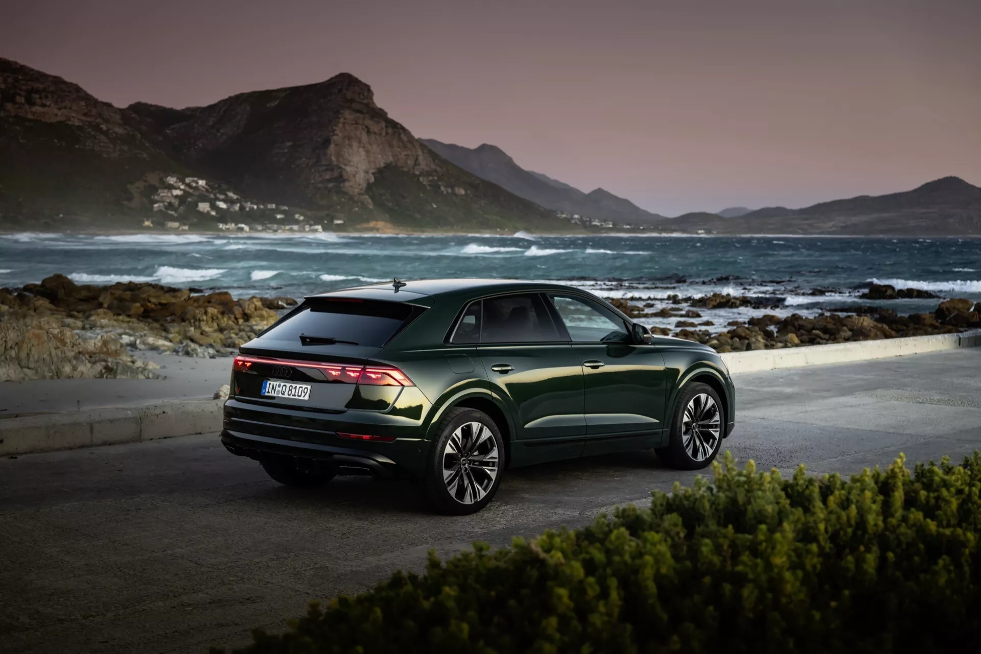 Audi Q8 2023 (4M-Facelift) garée dans un lieu pittoresque au bord de la mer à l'aube ou au crépuscule, présentant sa vue arrière trois quarts, finition vert métallique foncé, et jantes en alliage à multiples branches contre un fond montagneux serein