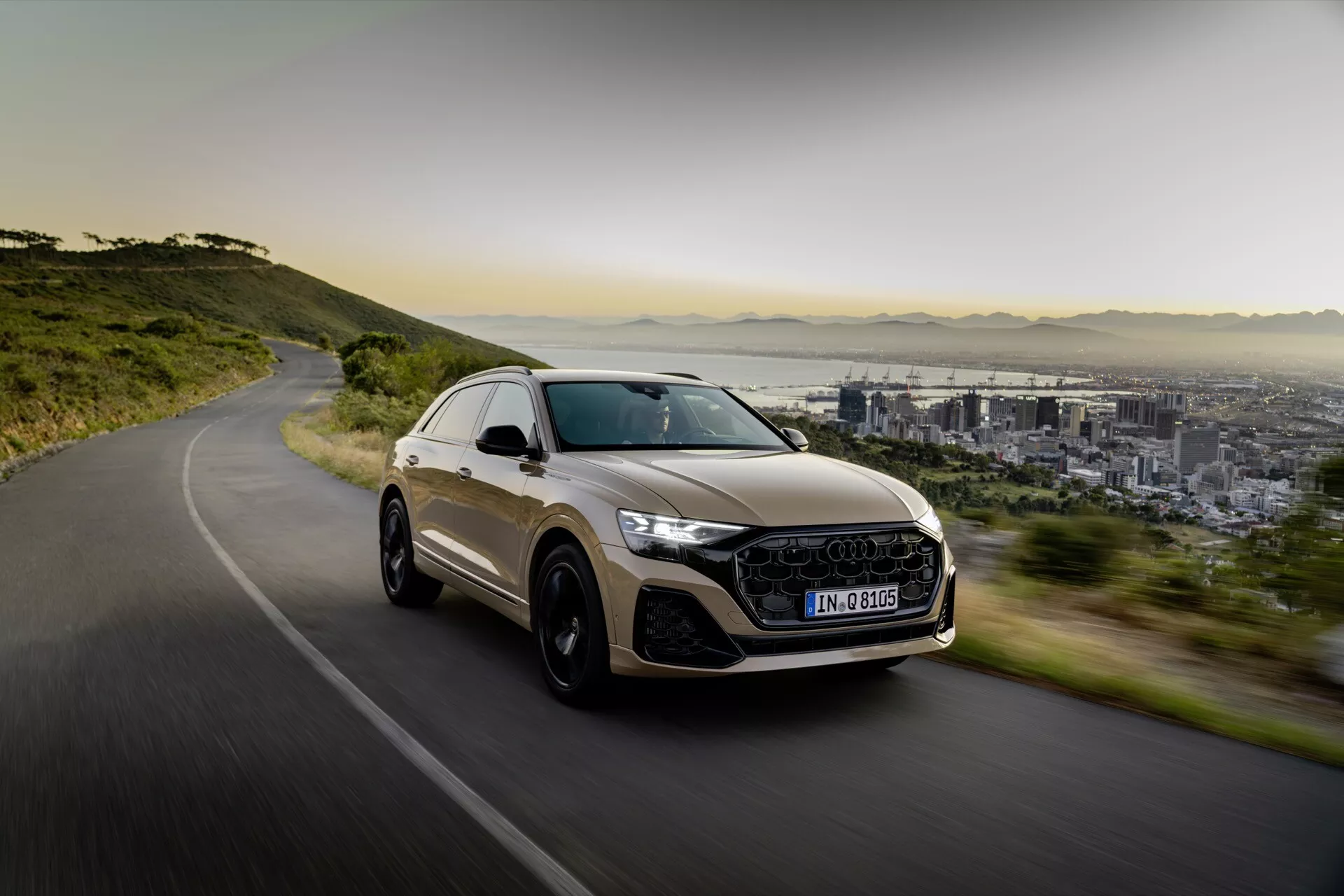 SUV crossover de luxe Audi Q8 2023 (4M-Facelift) en mouvement sur une route sinueuse, mettant en avant un design sportif, des phares LED, de grandes roues en alliage et un toit ouvrant panoramique, avec en arrière-plan la ville et les montagnes dans une lumière douce du matin ou de l'après-midi.