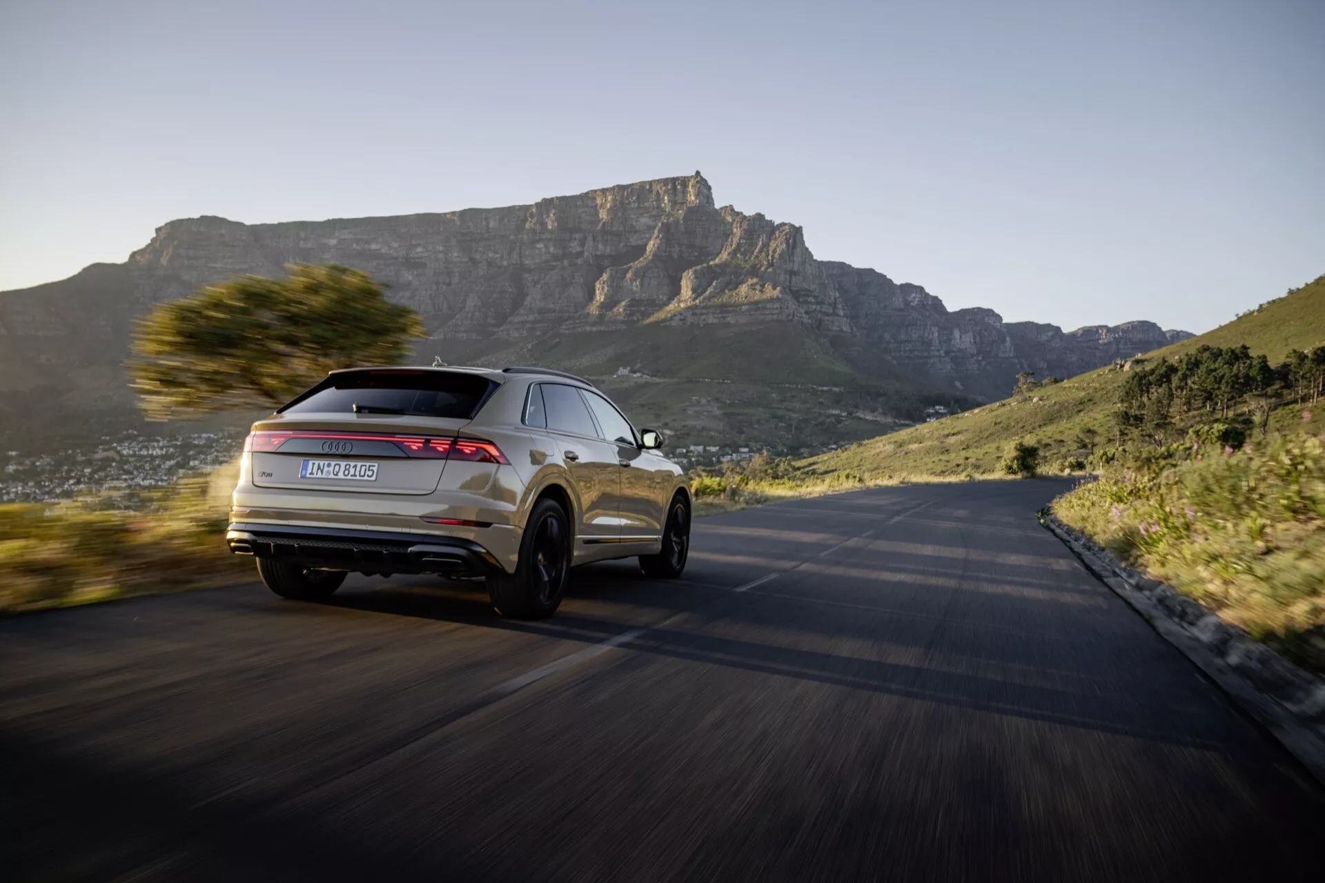 Modèle Audi Q8 4M facelift 2023 en mouvement dynamique sur une route courbée, mettant en avant son design moderne de SUV et fonctionnalités avancées depuis une perspective arrière de trois quarts, avec un fond de paysage montagneux au coucher du soleil