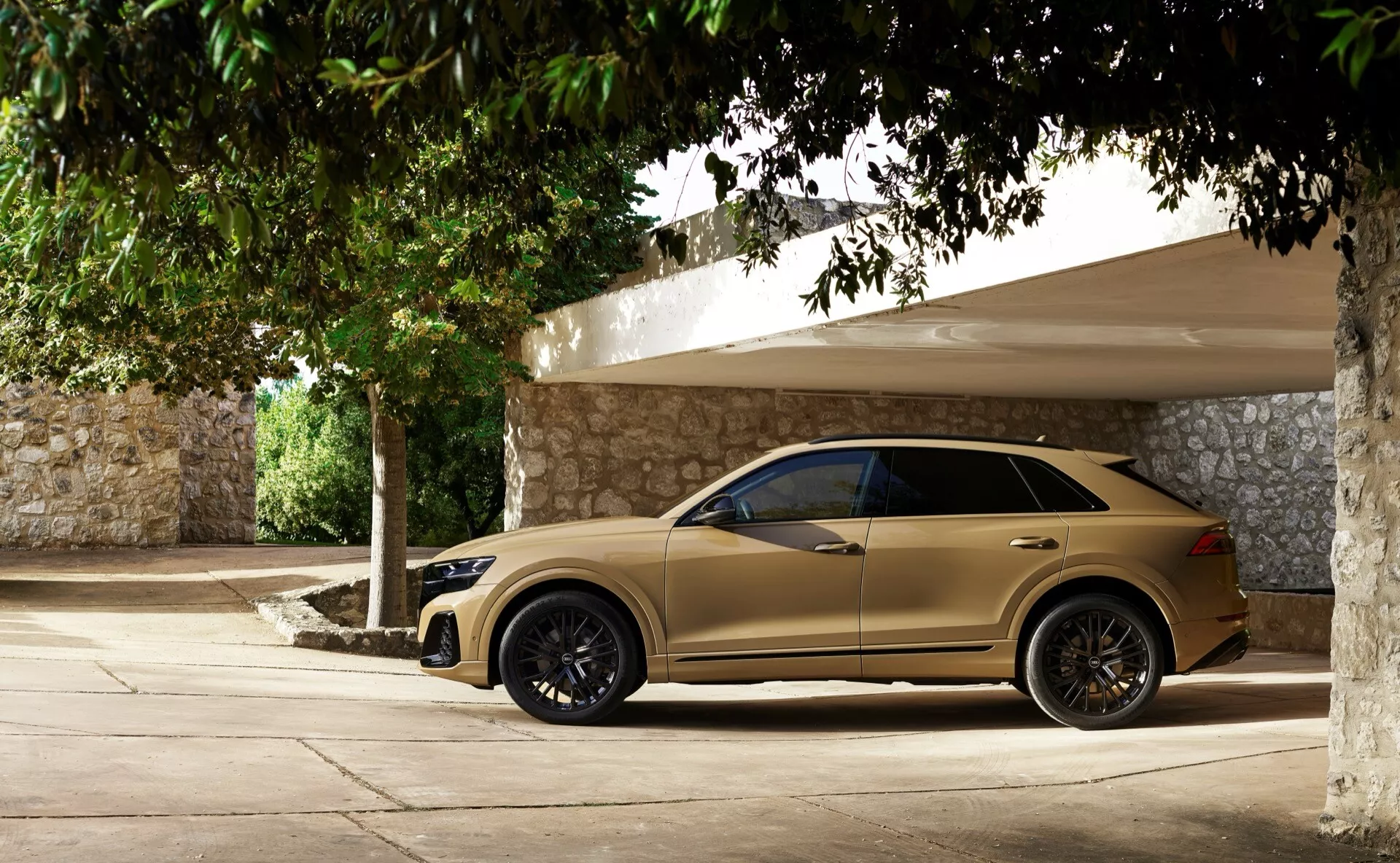 Audi Q8 2023 (4M-Facelift) en beige/doré, présentant une vue latérale complète avec jantes en alliage foncées, miroirs avec clignotants intégrés, vitres teintées, poignées de porte affleurantes et ligne de toit inclinée, garée à l'ombre lors d'une journée ensoleillée