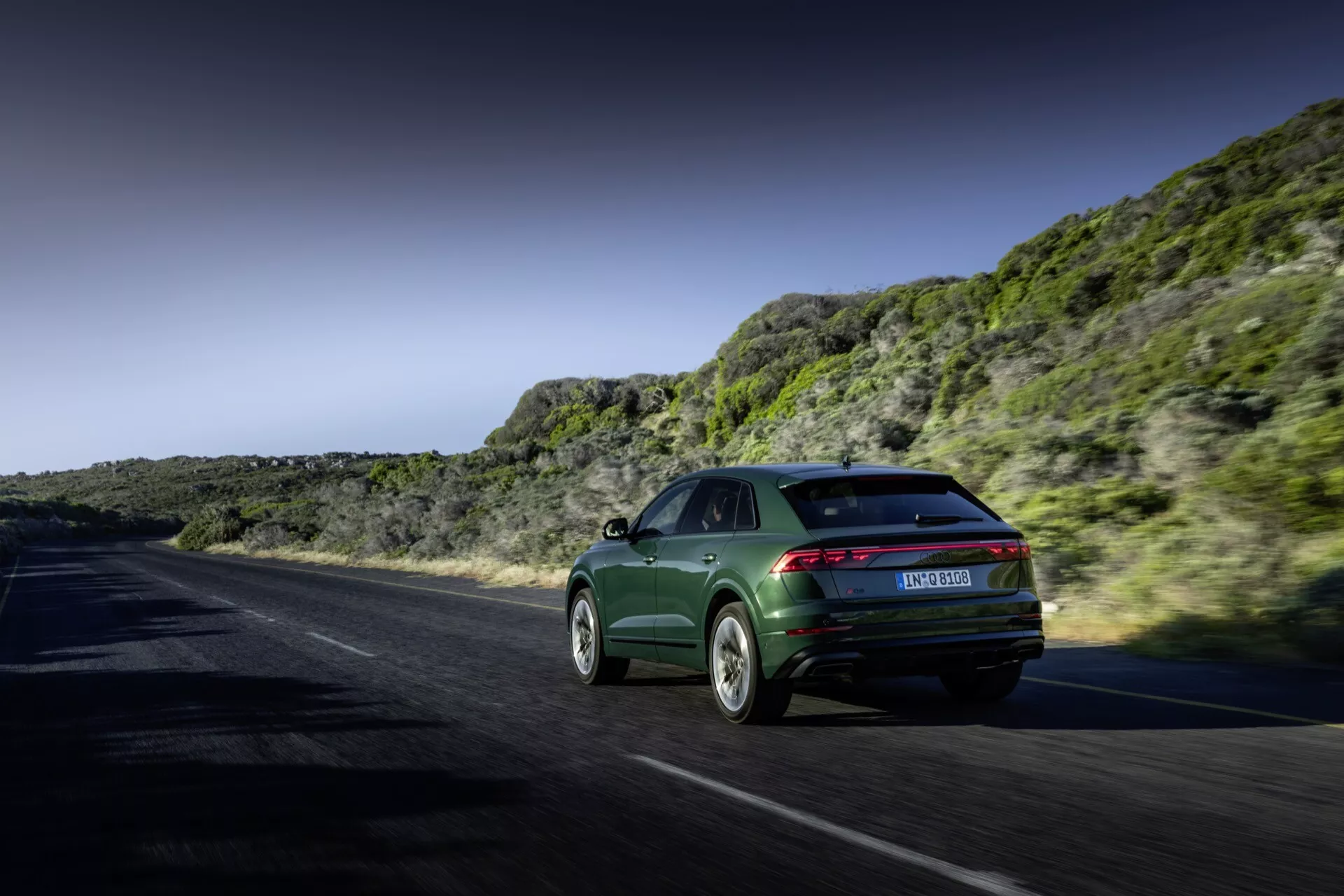 Audi Q8 2023 (4M-Facelift) en mouvement dynamique sur une route côtière ou montagneuse, présentant son style sportback, ses feux arrière élégants et ses roues en alliage sophistiquées contre un paysage dramatique