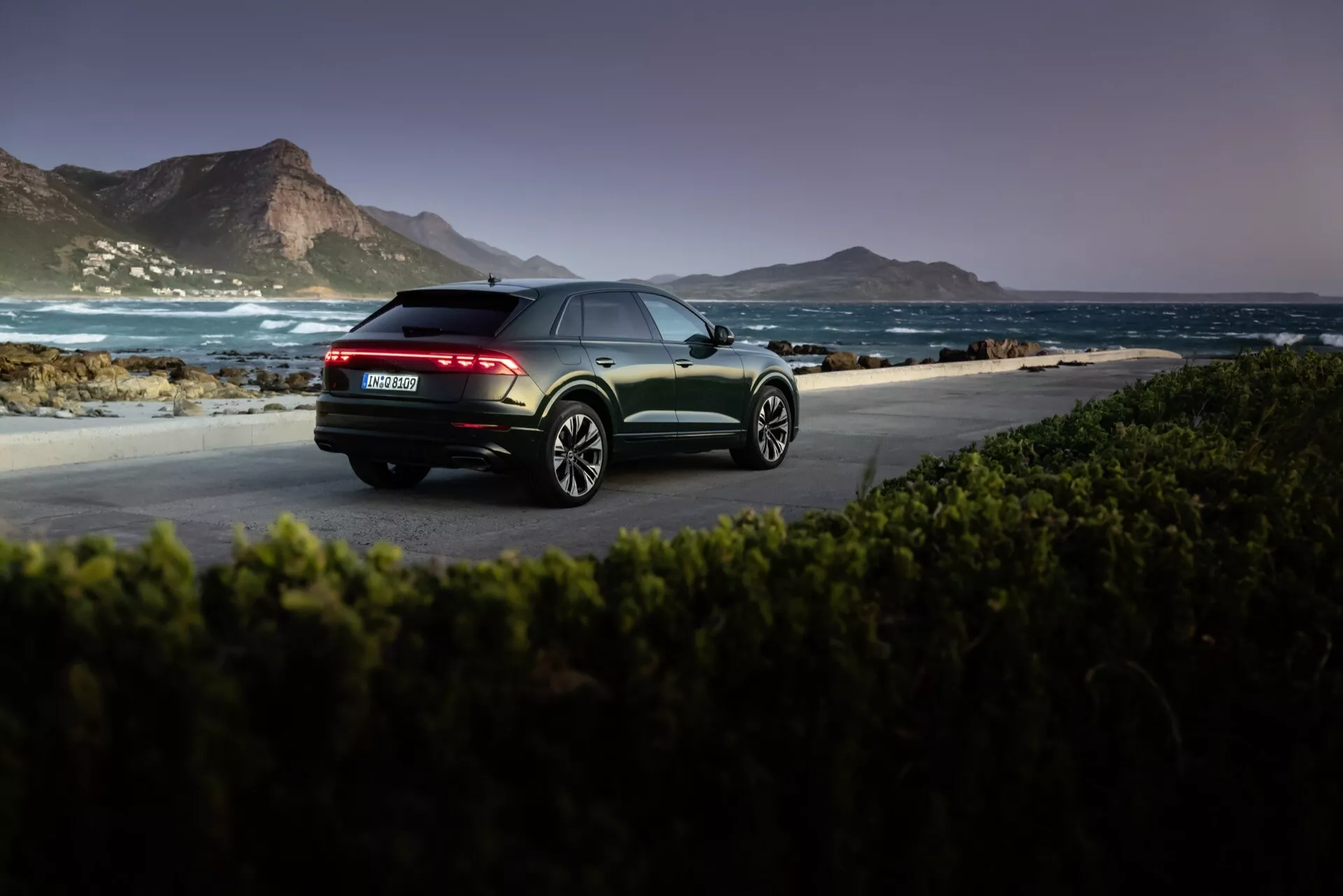 Audi Q8 4M-Facelift 2023 en finition métallique capturée à l'aube ou au crépuscule dans un cadre côtier naturel, mettant en lumière la vue arrière trois quarts avec feux arrière LED, double échappement et jantes en alliage à multiples branches.