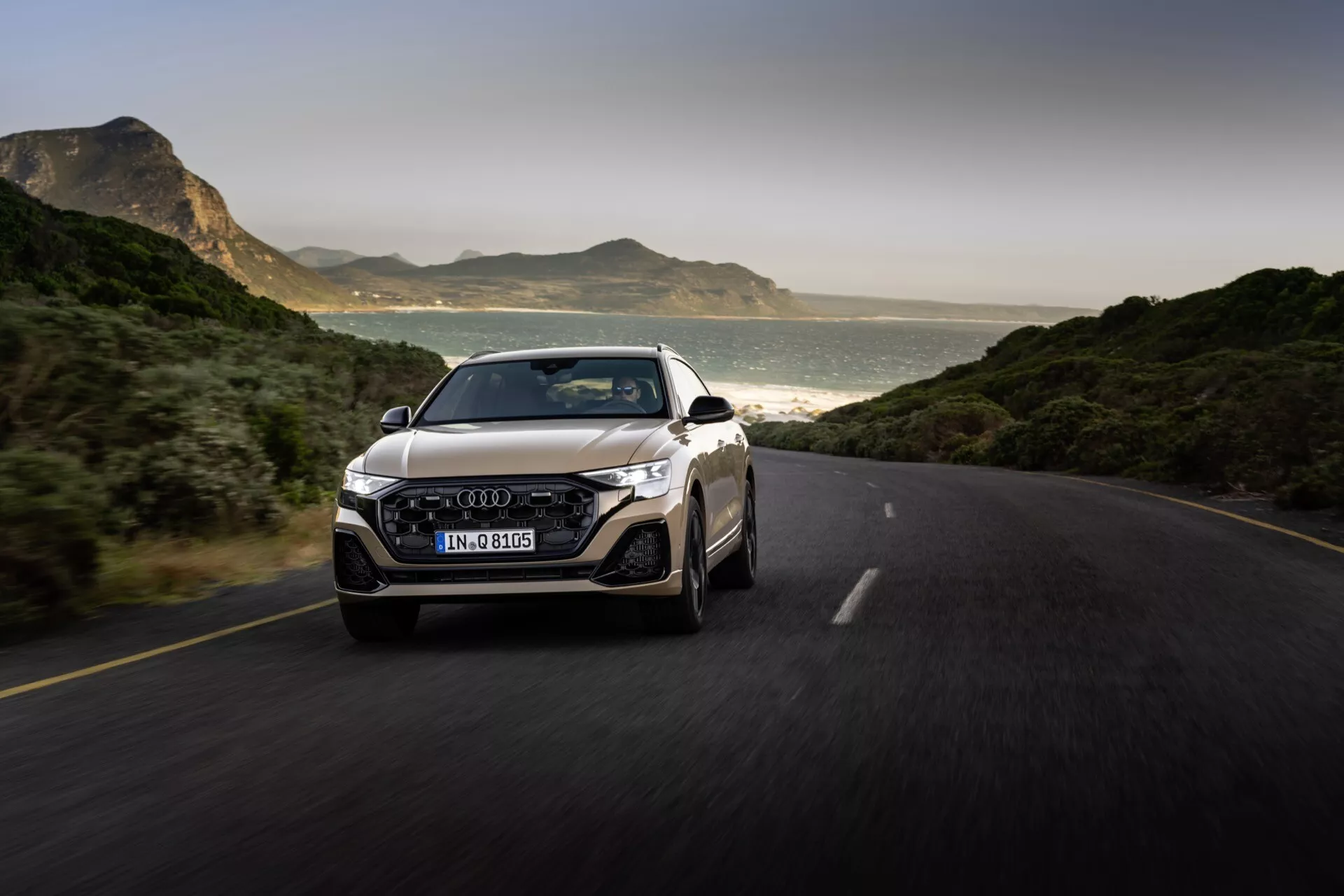 Audi Q8 2023 (4M-Facelift) capturée en mouvement sur une route pavée, mettant en valeur son design dynamique depuis une vue avant de trois quarts avec la grille, les phares LED, le pare-chocs, et l'emblème sur la grille, sur un fond de montagnes et de côte éclairé naturellement
