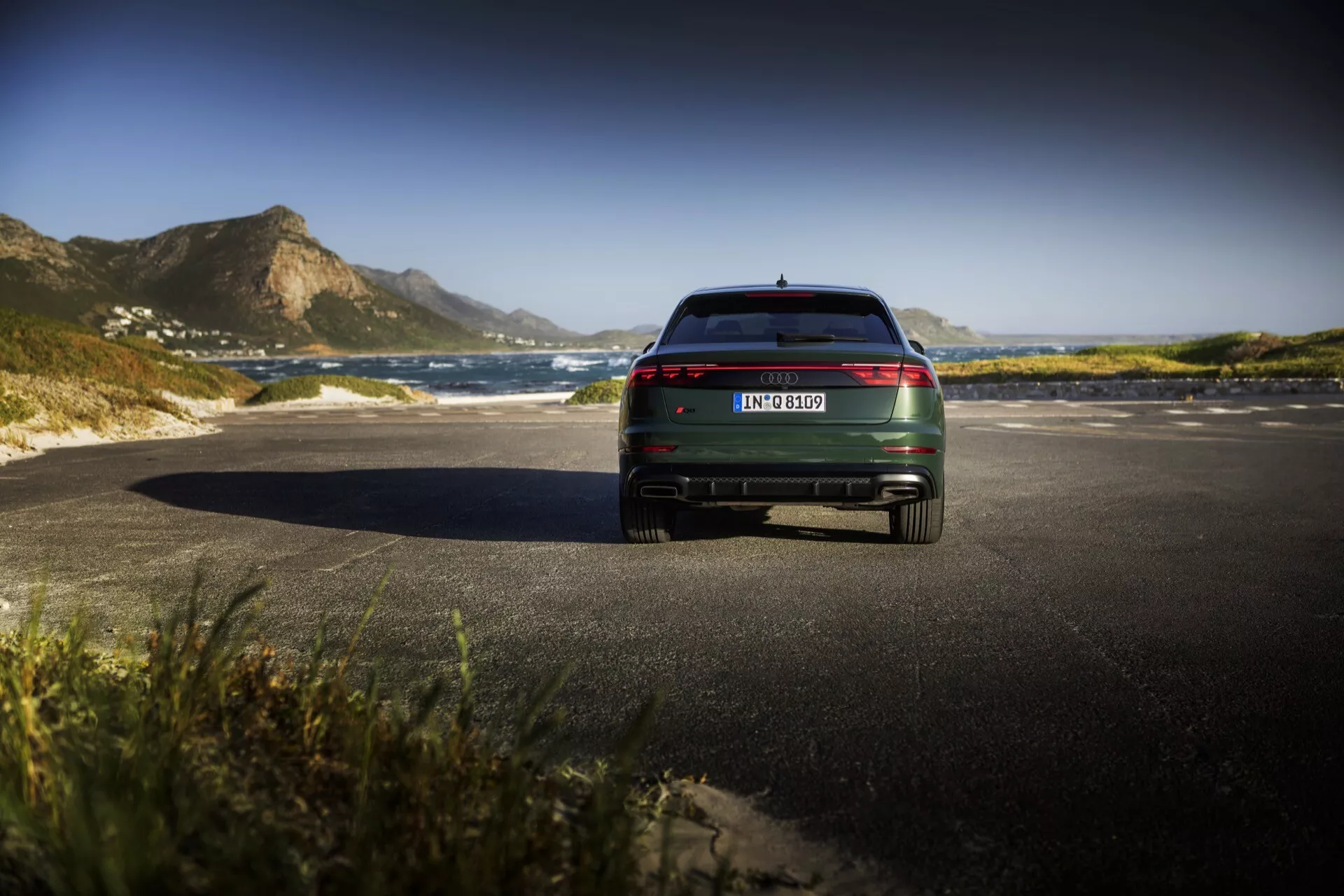 Vue arrière du modèle Audi Q8 4M Facelift 2023, montrant les feux arrière LED, les accents chromés et le double échappement sur une route côtière pittoresque