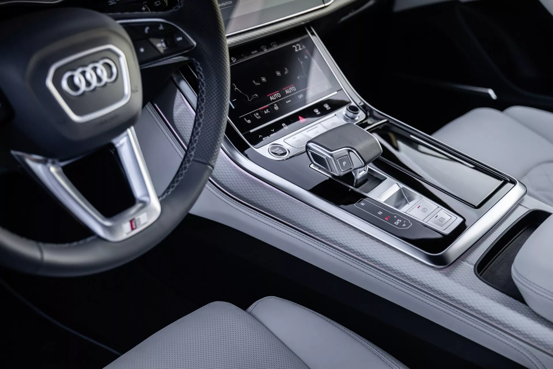 Vue rapprochée de la console centrale et d'une partie du volant de l'Audi Q8 2023 (4M-Facelift) depuis le côté passager, mettant en lumière les boutons du volant multifonctions, l'écran tactile central de l'infodivertissement, la zone de contrôle climatique et le levier de vitesse moderne dans une finition intérieure haut de gamme.