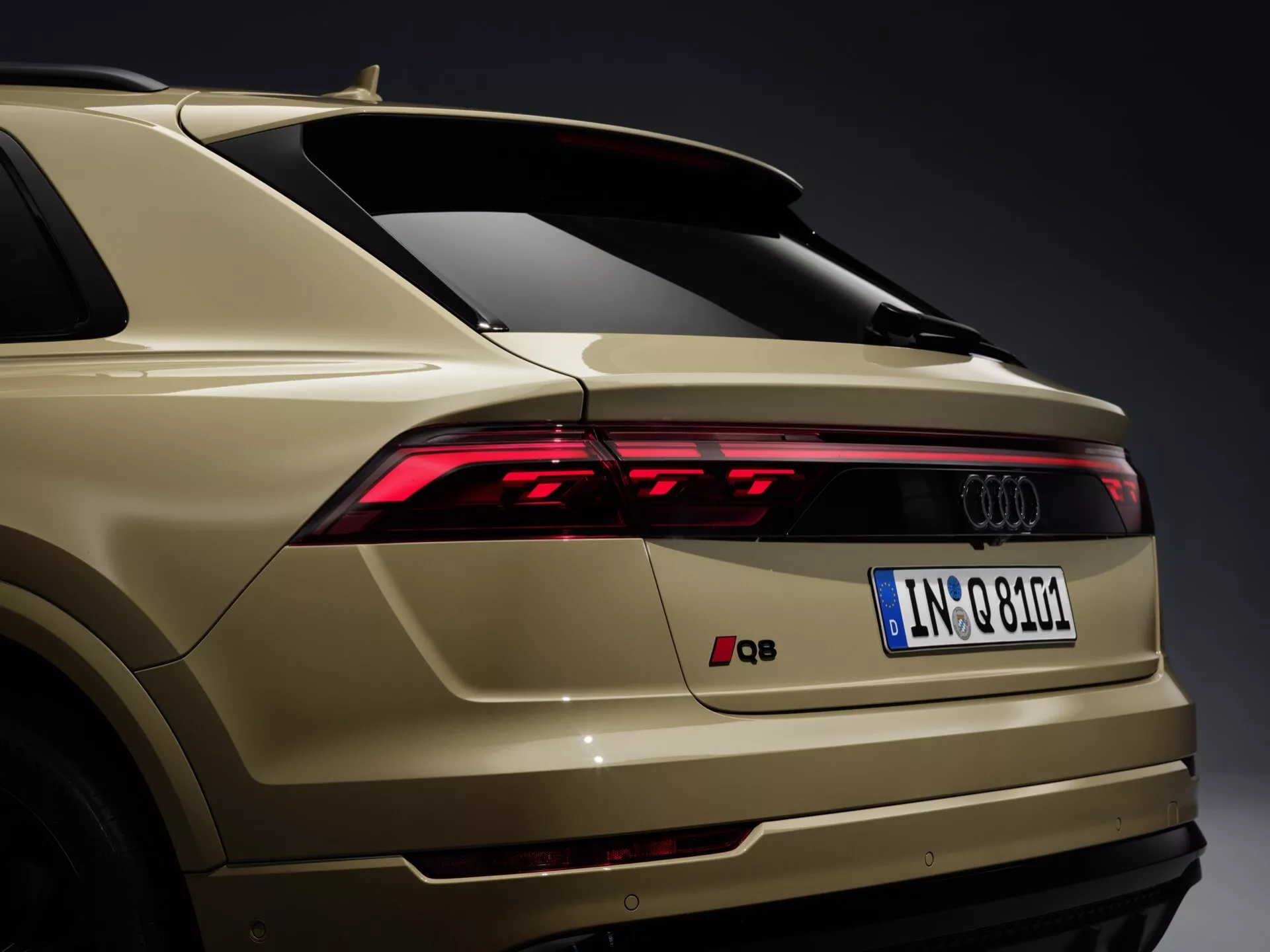 Vue arrière trois quarts de l'Audi Q8 4M-Facelift 2023 montrant des feux arrière LED, pare-chocs arrière élégant avec capteurs intégrés, branding Audi, plaque d'immatriculation européenne, spoiler arrière, finition peinture or-beige et antenne aileron de requin sur fond gris neutre.
