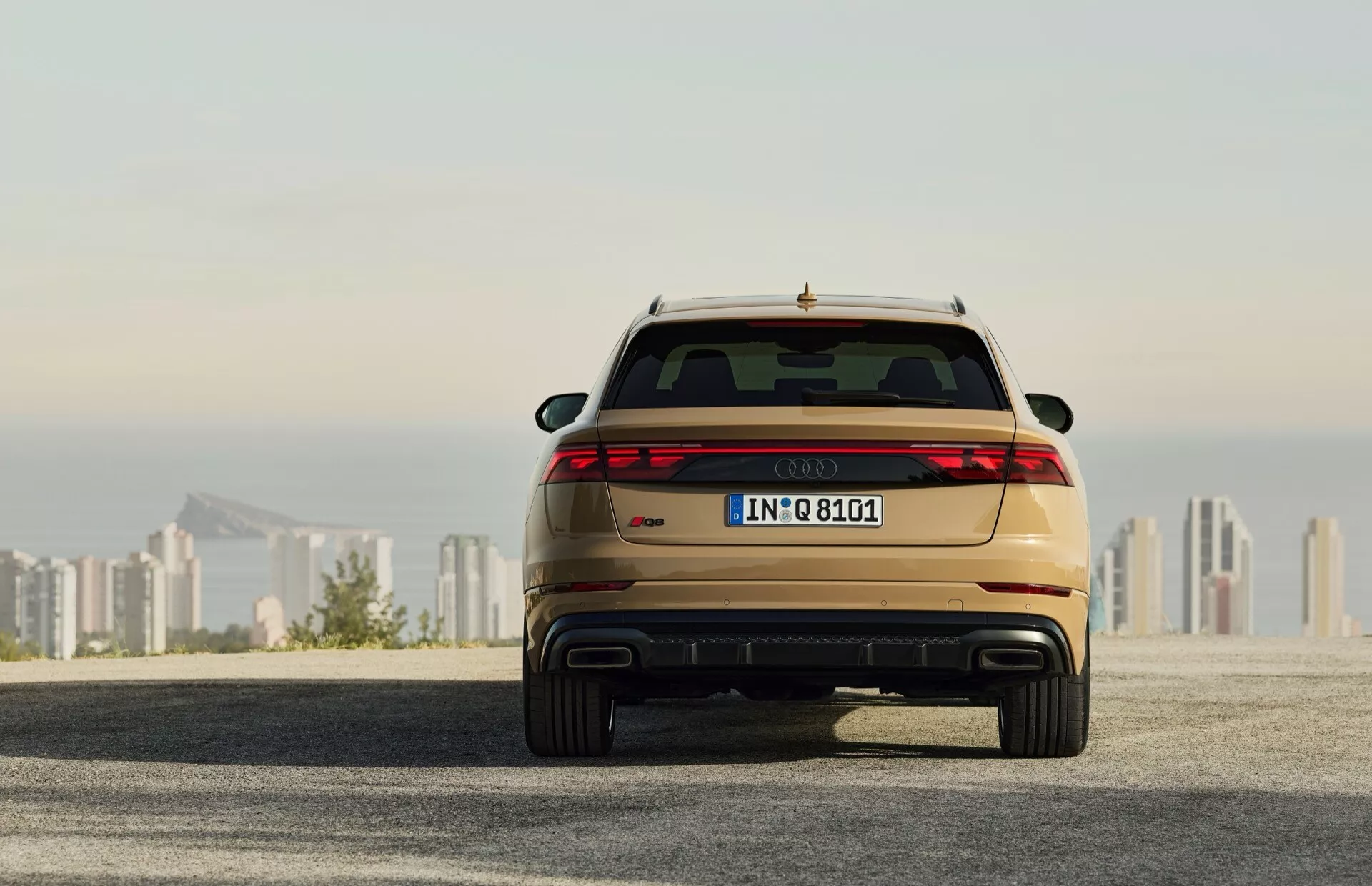 Vue arrière de l'Audi Q8 2023 (4M-Facelift) montrant des feux arrière LED, un double échappement et une peinture métallique, garée sur une surface en asphalte avec l'horizon de la ville en arrière-plan au lever/coucher du soleil