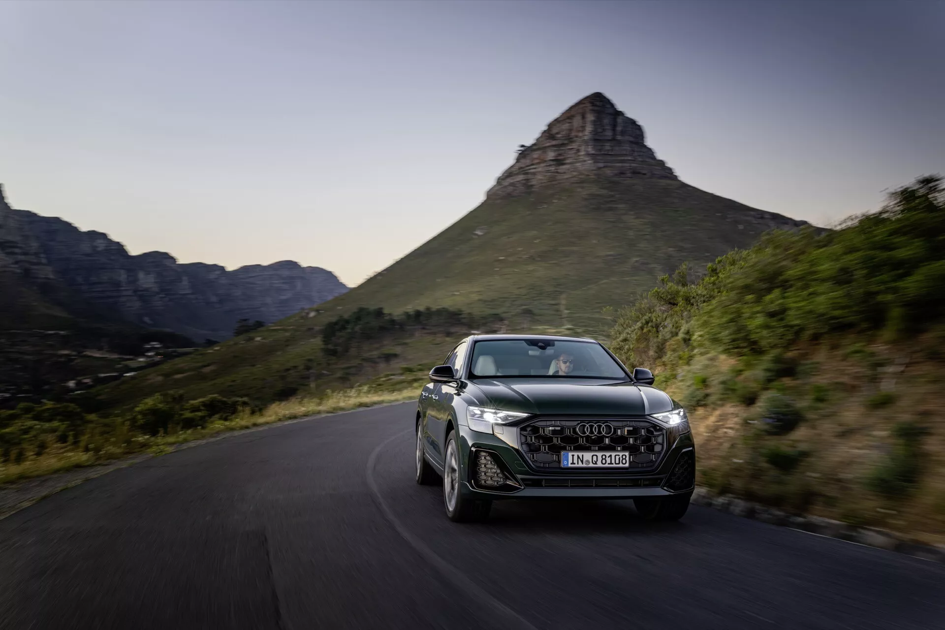 Audi Q8 2023 (4M-Facelift) en mouvement dynamique sur une route sinueuse pittoresque, présentant son design aérodynamique, phares LED, jantes en alliage distinctives et prises d'air intégrées sur fond de montagne pittoresque en fin d'après-midi