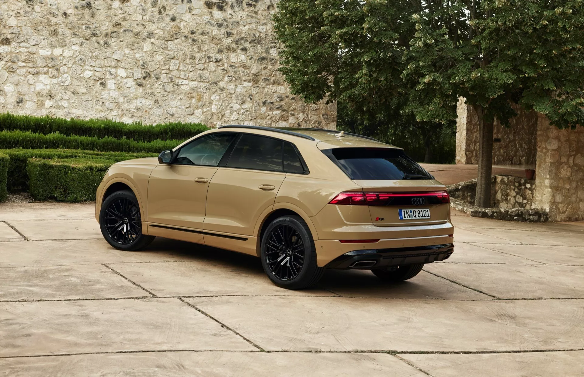 Audi Q8 2023 (4M-Facelift) exposée dans une vue arrière aux trois quarts présentant de grandes roues en alliage multi-branches noires, peinture métallisée or-beige, feux arrière LED horizontaux allongés, becquet arrière et système d'échappement double devant un mur en pierre historique