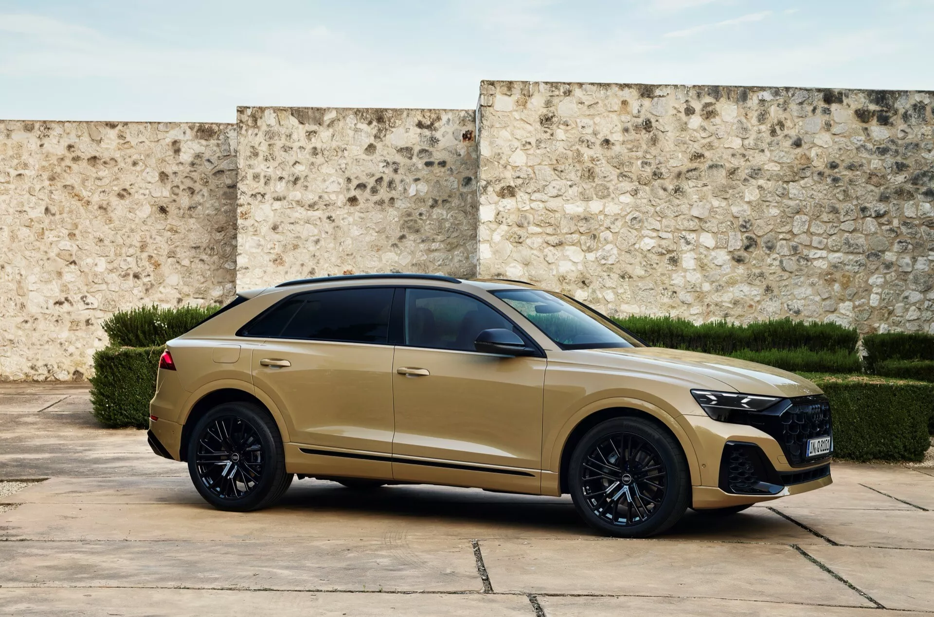 Vue de trois quarts avant de l'Audi Q8 2023 (4M-Facelift) moderne en finition mate dorée ou beige, présentant une large grille à motif en nid d'abeille, des phares LED, un pare-chocs avant sportif, des jantes en alliage noir, des pneus à profil bas, des poignées de porte affleurantes, des vitres teintées foncées, une ligne de toit fuyante style coupé, une ligne d'épaule musclée se rejoignant avec des feux arrière angulaires, et un aileron de toit, située devant un mur texturé évoquant un cadre historique ou contemporain.