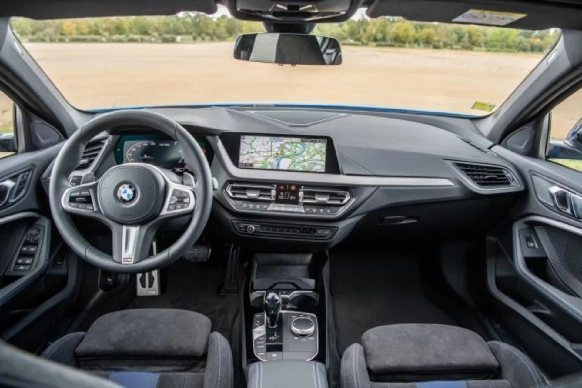 Vue intérieure de la BMW Série 1 M Performance 2019 depuis le siège arrière montrant le tableau de bord, la console centrale et le volant avec commandes intégrées, système d'infotainment tactile affichant la navigation GPS, aérations, commandes de climatisation, levier de vitesse automatique, molette rotative et pavé tactile sur la console centrale, sellerie premium et habitacle clair et bien éclairé
