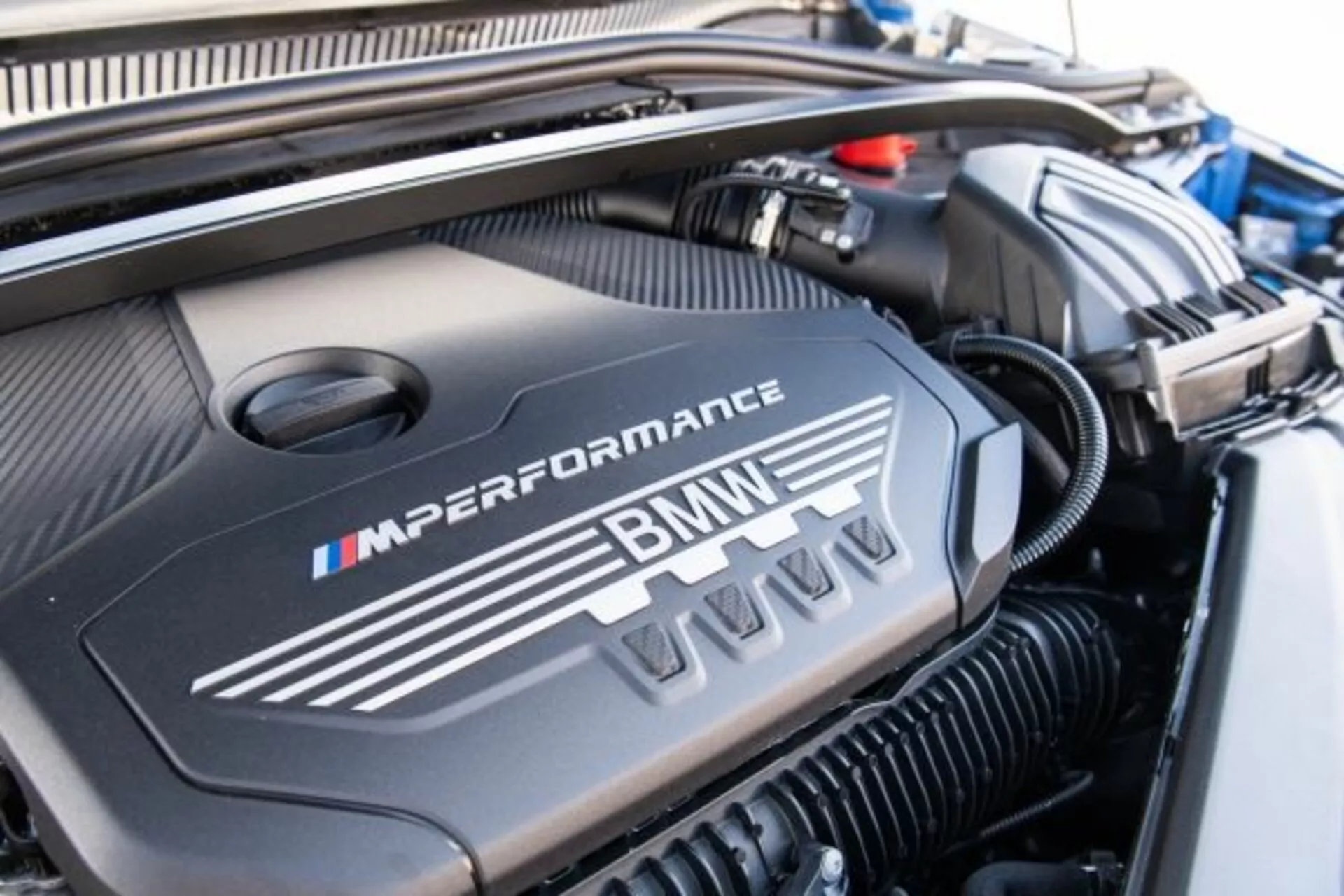 Image en gros plan du compartiment moteur d'une BMW Série 1 M Performance 2019, mettant en vedette le couvercle du moteur orné du logo M dans ses couleurs emblématiques bleu, violet et rouge, soulignant la conception sportive et haute performance.